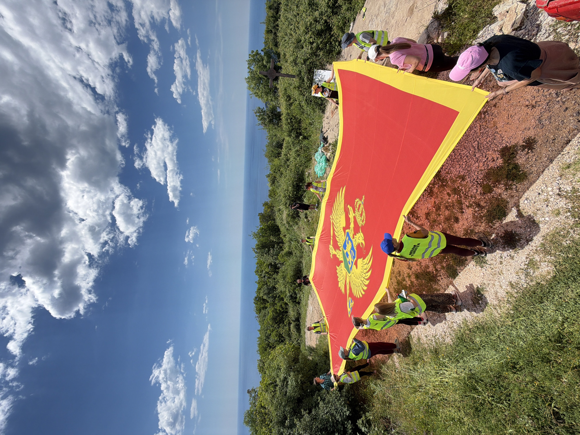 The image shows a group of people holding a large Montenegrin flag on what appears to be a hillside. The flag is predominantly red and yellow, featuring the Montenegrin coat of arms in the center. The people holding the flag are wearing casual clothing, some with safety vests. The location appears to be near a coastal area, as the sky with scattered clouds and a body of water are visible in the background. There's a wooden cross on the hillside. Based on the geographical features and flag, the location is likely near Budva, Montenegro. The people are gathered on rocky and grassy terrain.