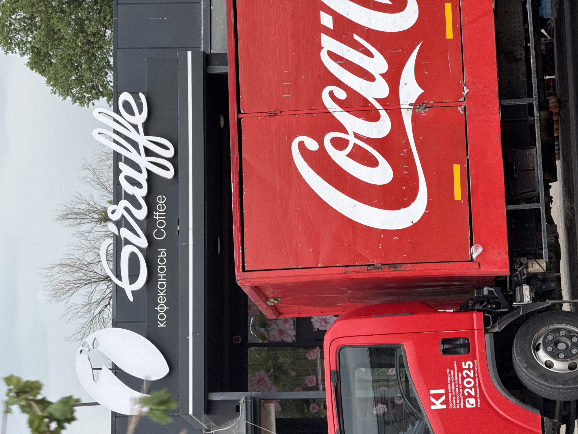 A red Coca-Cola delivery truck is parked in front of a cafe named "Giraffe". The cafe has a black facade with white lettering. The truck has a large Coca-Cola logo on its side. The cafe's windows are decorated with floral patterns. There are trees in the background, suggesting an outdoor setting. The sky is overcast.