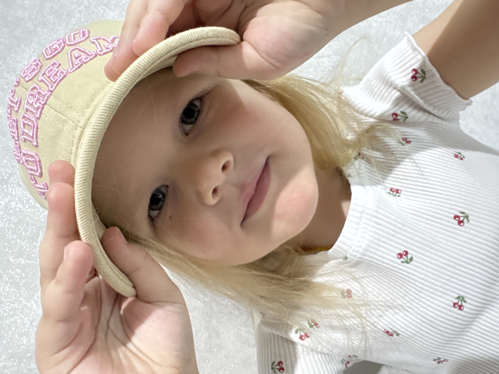 A young, fair-skinned child with blonde hair is holding a beige cap with pink lettering on the brim. The child's face is angled towards the camera. They are wearing a white, ribbed shirt with a cherry pattern. The background appears to be a white, textured surface. The location of the scene is Gornyi (Dvurechenskoe S/P), Russia.