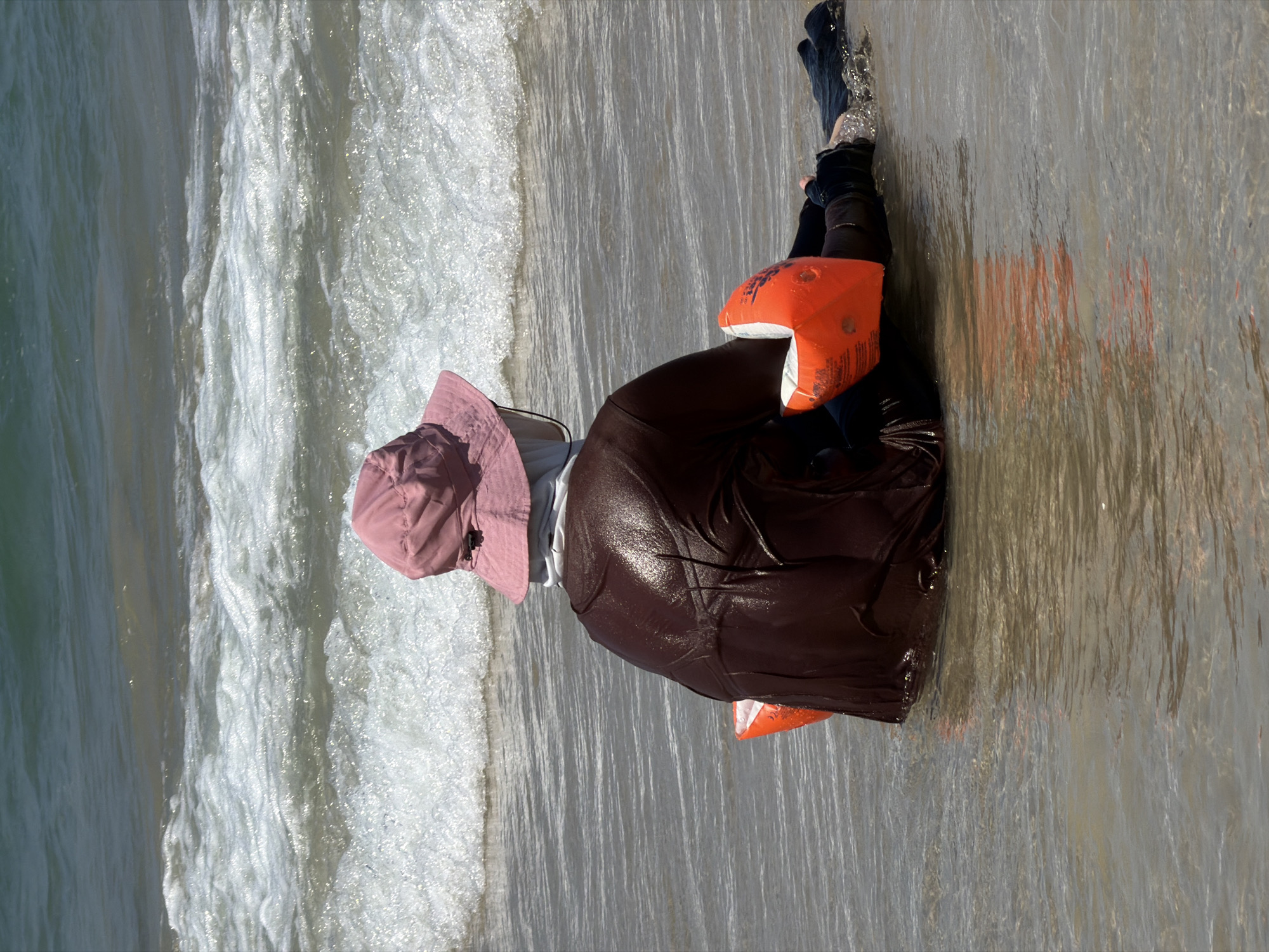 A person wearing a pink sun hat and a dark wet garment with orange floatation devices on their arms sits in shallow water on a beach in Bat Yam, Israel. The person is facing away from the camera, towards the ocean. The water is clear and has small waves breaking. The background consists of the ocean and a bright, sunny sky.
