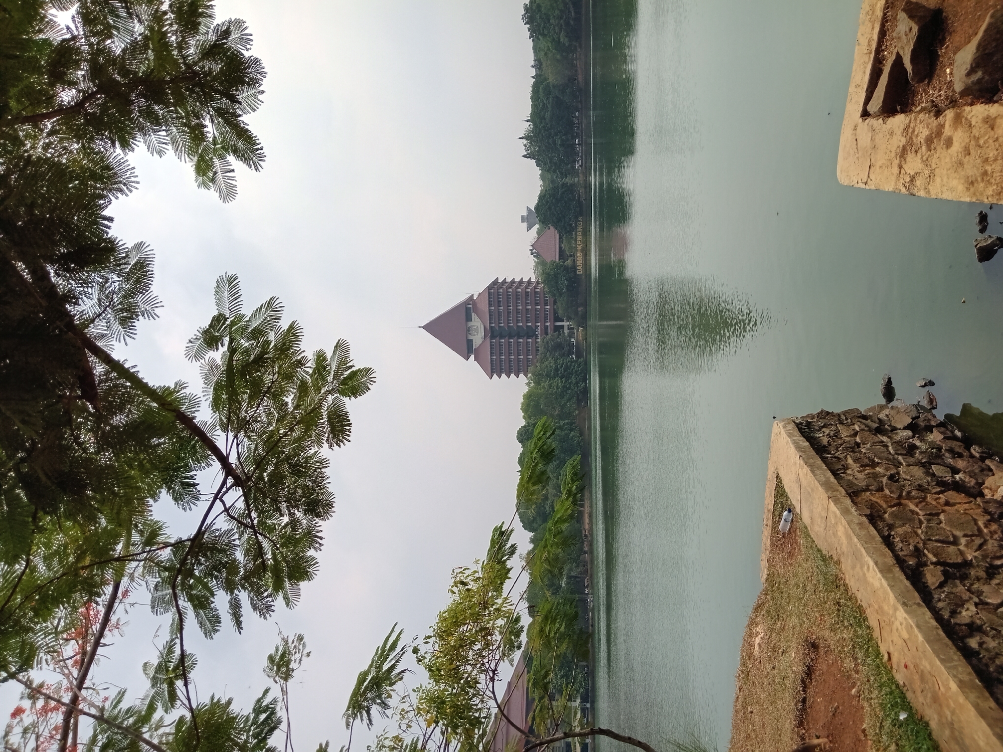 A calm, light green lake is visible in the foreground, reflecting the overcast sky. The lake's edge is bordered by a concrete and stone retaining wall. Beyond the lake, a cluster of trees line the opposite shore, with a prominent, multi-story, pyramid-roofed building situated centrally amongst them. To the left of this building, a smaller structure with a similar architectural style is partially visible, and further to the left, a water tower-like structure stands. The name "DANAU KENANGA" is visible on a sign between the smaller building and the water tower. The sky is mostly clear, with some wispy clouds. Lush green foliage from trees overhangs the top of the frame. The scene is a tranquil outdoor setting, likely a park or campus area.
