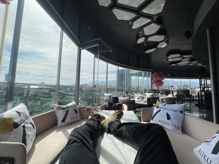 The image displays the interior of a modern, upscale rooftop bar or lounge, identified as the Roku Gin Garden at the InterContinental Sofia, located in Sofia, Bulgaria. The perspective is from a low vantage point, focused on a person's lower body.
In the immediate foreground, a person's legs are visible, extended onto a low, rectangular table with a white marble-patterned surface. The individual wears dark trousers and light brown, possibly suede, boots. They are seated on a light beige upholstered banquette. Two white cushions are prominent on the banquette, each printed with black Japanese-style brushstrokes and the text "ROKU GIN."
The lounge features extensive floor-to-ceiling glass windows that curve around the building, offering a panoramic view of the Sofia cityscape under a bright, partly cloudy sky. Distant mountains, consistent with Vitosha Mountain, are visible on the horizon. The city below consists of various buildings and rooftops.
Within the midground of the lounge, multiple seating arrangements are present, including light-colored upholstered sofas and chairs, alongside dark-patterned low tables. A striking artificial pink cherry blossom tree acts as a central decorative element. Further into the lounge, additional patrons are faintly discernible seated at tables.
The ceiling is dark, likely black, and is adorned with multiple geometric, hexagonal light fixtures composed of white translucent panels, some accented with small pink floral details. The floor surface appears dark and reflective. The overall scene depicts a relaxed, high-elevation urban environment during daylight hours.