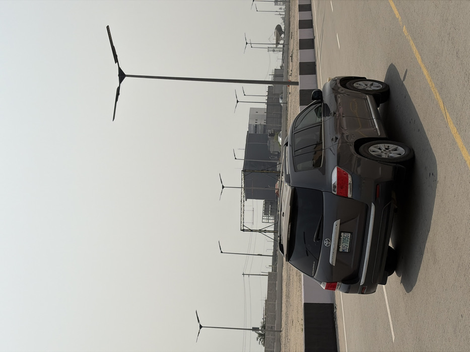 A dark grey Toyota Highlander SUV, bearing Nigerian license plate LUS-7056T, is depicted on an asphalt road in Alaguntan, Nigeria. The vehicle is viewed from its rear-right, positioned in the right-most lane, moving away from the perspective. The road surface features yellow lane markings and is flanked by a black and off-white striped concrete median barrier to the left. Numerous modern streetlights with flat, three-pronged LED fixtures line both sides of the thoroughfare, receding into the hazy, light grey sky. The background reveals an industrial area comprising several dark-colored industrial buildings, including a large structure and a distant, circular, dish-like apparatus on the far right. Sparse vegetation is visible behind the median barrier. No individuals are present in the scene.