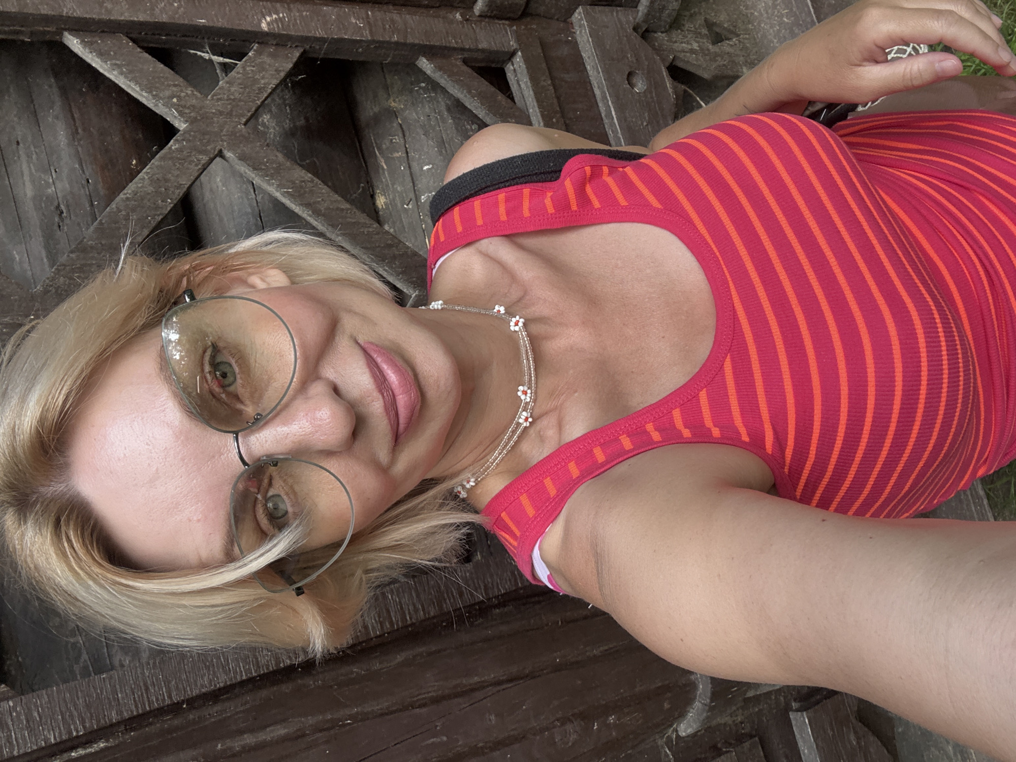 A woman is lying down on a wooden bench. She is wearing a pink and orange striped tank top and a necklace with small white and red flowers. She has blonde hair and is wearing large, round glasses. Her left arm is resting on the bench next to her, and her right arm is bent, with her hand resting on her stomach. Her head is tilted back, and she is looking towards the camera with a slight smile.
The setting appears to be outdoors, possibly in a park or garden, given the presence of grass visible in the background and the rustic wooden bench. The lighting suggests it could be daytime, with natural light illuminating the scene. The overall mood seems relaxed and casual. There is no visible text in the image.