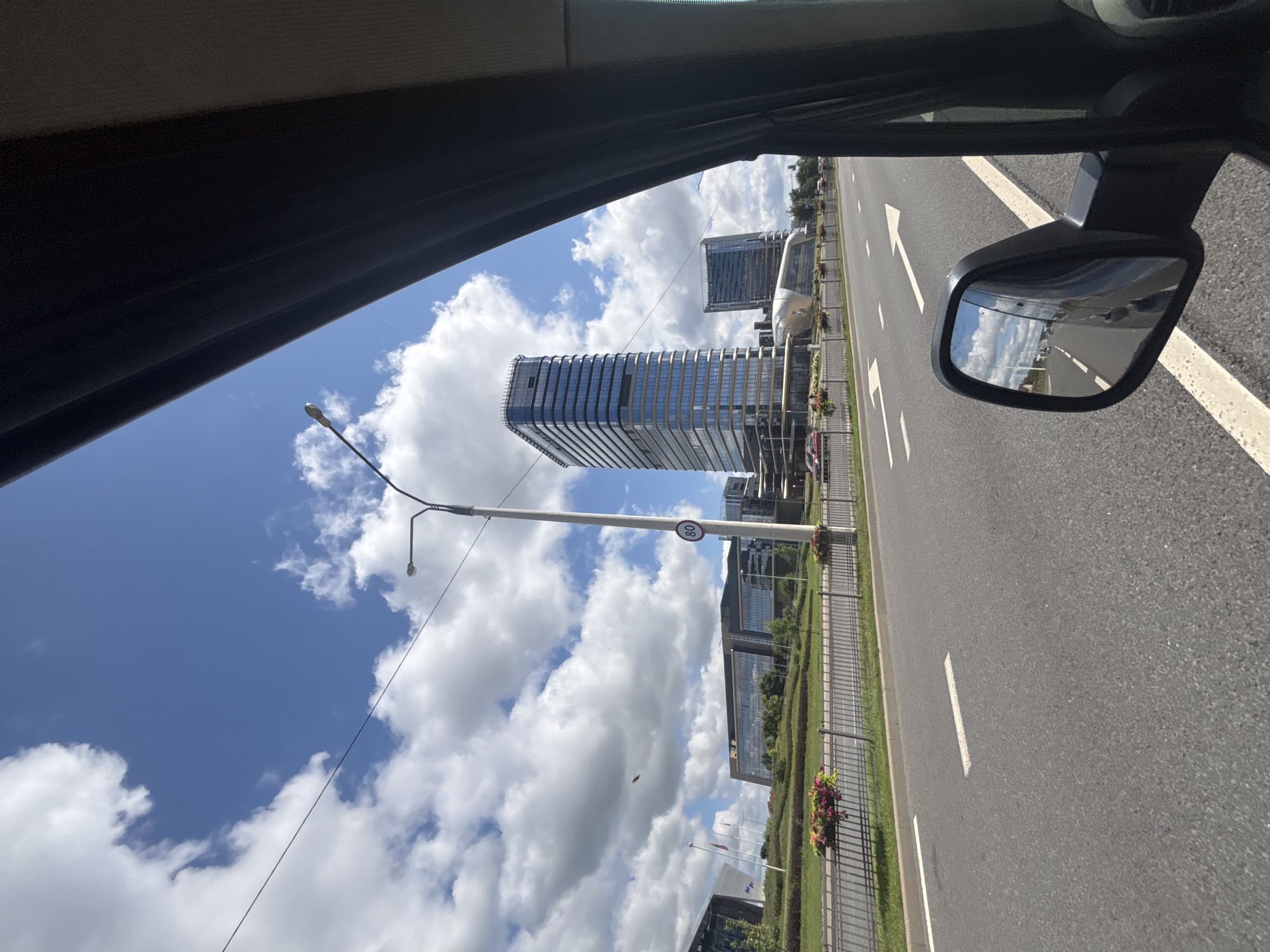 The image depicts a view from inside a vehicle, looking out towards a modern cityscape in Minsk, Belarus, on a clear day with scattered clouds. A multi-lane asphalt road dominates the foreground, with white lane markings and directional arrows visible. To the left of the road, a black metal fence separates the road from a neatly manicured grassy area with flower beds. Several contemporary glass-fronted buildings, including a tall skyscraper, are visible in the background, suggesting a business or commercial district. A street lamp with a speed limit sign (80) is positioned along the roadside. The side-view mirror of the vehicle reflects the road ahead and the sky, showing the continuous flow of traffic and the bright, cloudy sky. The overall scene conveys a sense of urban development and everyday life in motion.