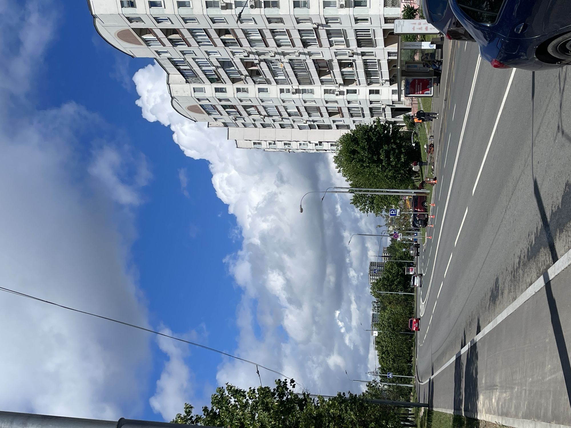 The image depicts a street scene in Moscow, Russia, characterized by a wide asphalt road with white lane markings. A modern, multi-story apartment building with numerous windows and balconies dominates the right side of the frame. Trees line the left side of the road, with a sidewalk and a grassy verge. A few cars are visible on the road, including a red car in the distance and a dark blue car in the foreground. Several traffic cones are placed along the road, suggesting ongoing construction or maintenance. A bus stop with a shelter and advertising posters is situated on the sidewalk near the apartment building. A few individuals are present near the bus stop and by the side of the road, some appearing to be workers. The sky is a bright blue with large, fluffy white clouds. The overall impression is a typical urban environment on a sunny day.