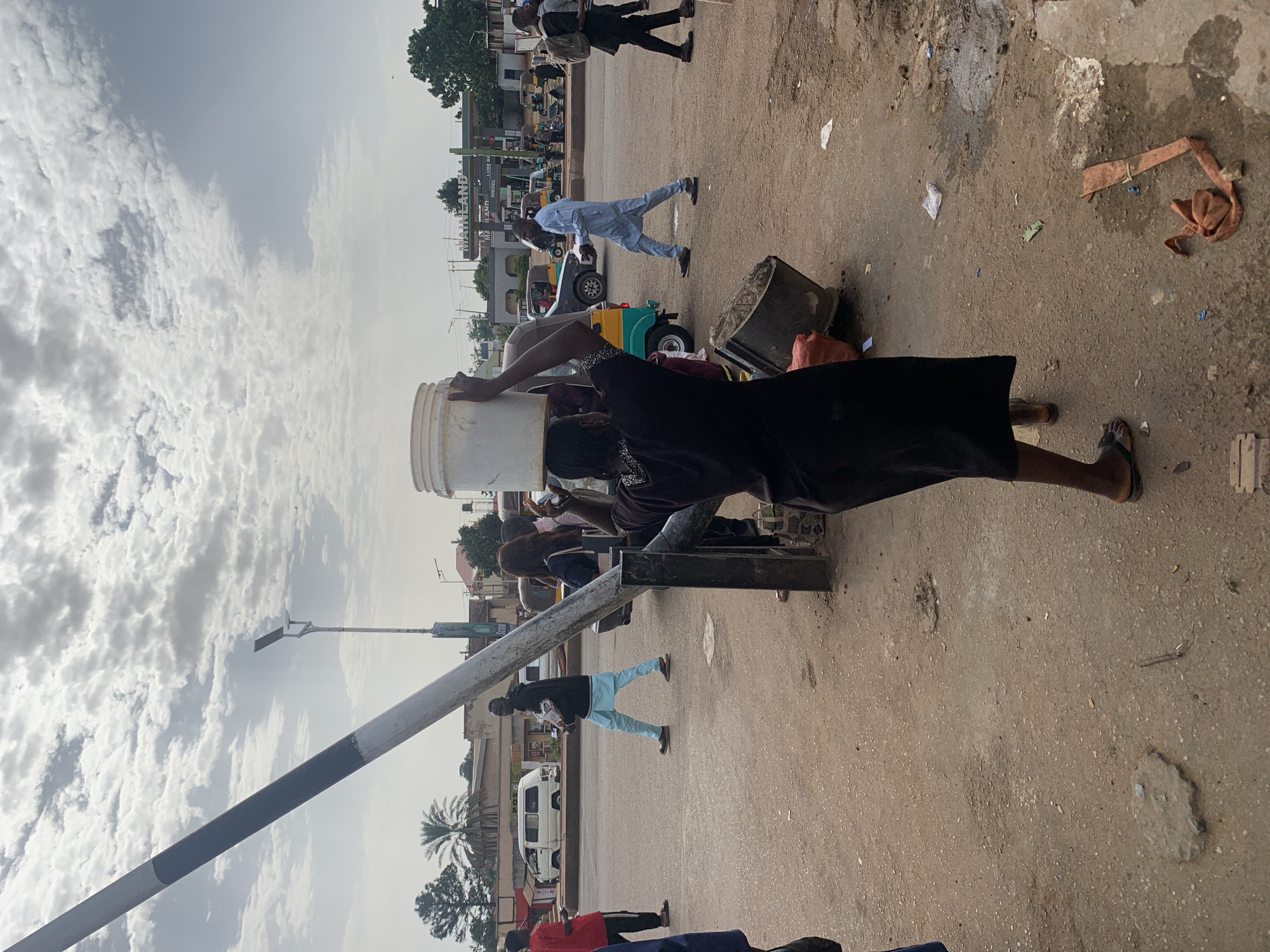 A person is walking on a dirt road, carrying a bucket of water on their head. The person is wearing a dark dress and sandals. The road is lined with buildings and trees. There are several vehicles on the road, including a white van, a blue car, and a yellow and green tuk-tuk. In the background, there is a gas station with "MAINLAND" written on it. The sky is cloudy. The location is Gura Top, Nigeria.