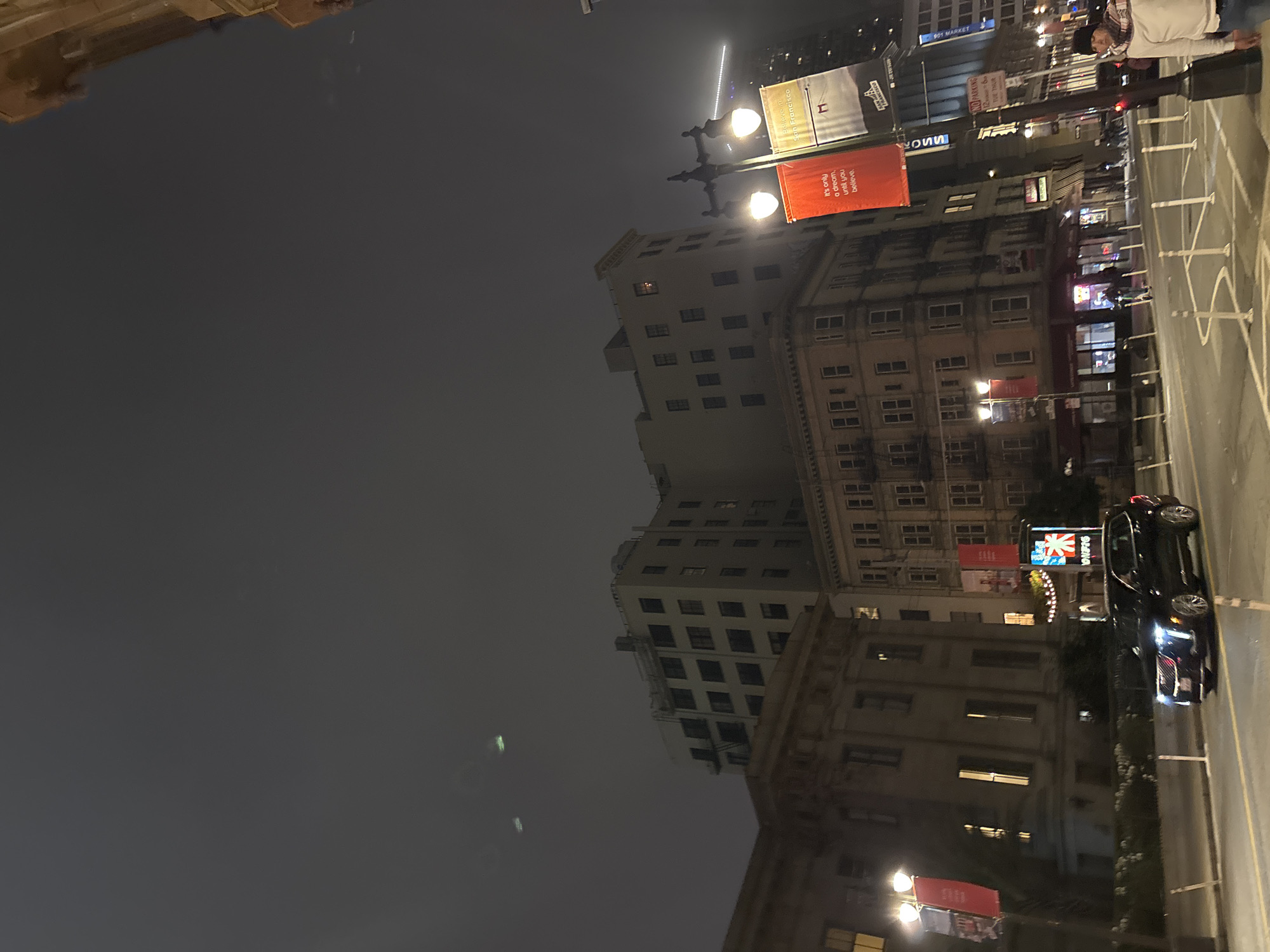 This is a nighttime street-level view of San Francisco, California. The sky is overcast and dark, with a few faint green lights visible in the upper portion of the image, possibly from aircraft or atmospheric phenomena. In the foreground, a black Cadillac SUV is driving on a wet street. To the left of the SUV, there is a sidewalk with bollards and planters. Across the street, there are several buildings of varying heights and architectural styles. One building on the left appears to be a light-colored, multi-story structure with a classical facade, possibly a government building or historical landmark. Adjacent to it is a taller, lighter-colored building with many windows, appearing to be an apartment or office building. Further to the right, a tan-colored building with a more ornate facade is visible, with numerous windows and balconies. Streetlights illuminate the scene, with two bright lamps visible on a lamppost in the mid-ground. Two banners hang from the lamppost; one is red and displays text, while the other is yellow and also has text and a graphic. A man in a white shirt and a patterned scarf is standing on the sidewalk to the far right of the image, looking towards the street. There are other people and vehicles present in the background, but they are less distinct due to the lighting conditions and distance. A sign with "NO PARKING" and other text is visible on a pole in the mid-ground on the right.