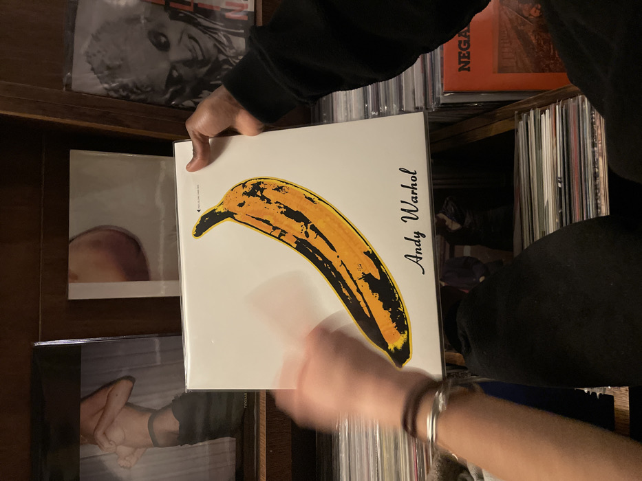 An overhead-angle shot captures two individuals interacting with a vinyl record inside a record store in Paris, France. The central object is a white album cover featuring Andy Warhol's yellow and black banana artwork, with "Andy Warhol" printed below. The record is enclosed in a clear plastic sleeve, bearing a small black text reading "PEEL SLOWLY AND SEE" in the top right corner. A dark-skinned right hand, partially covered by a black sleeve, grips the top right corner of the album. A lighter-skinned left hand, adorned with a silver bracelet and a brown cord bracelet, appears motion-blurred as it interacts with the left side of the album. Below the album, numerous other vinyl records are stacked horizontally on wooden shelves. In the background, above the central album, three distinct album covers are visible on an upper wooden shelf: a black and white image of a shirtless torso from the chest down on the left, a minimalist cover with a reddish-brown circular object on a light background in the center, and a black and white portrait of a person with textured hair and a wide grin on the right. The overall environment suggests a well-stocked record shop with wooden display fixtures.