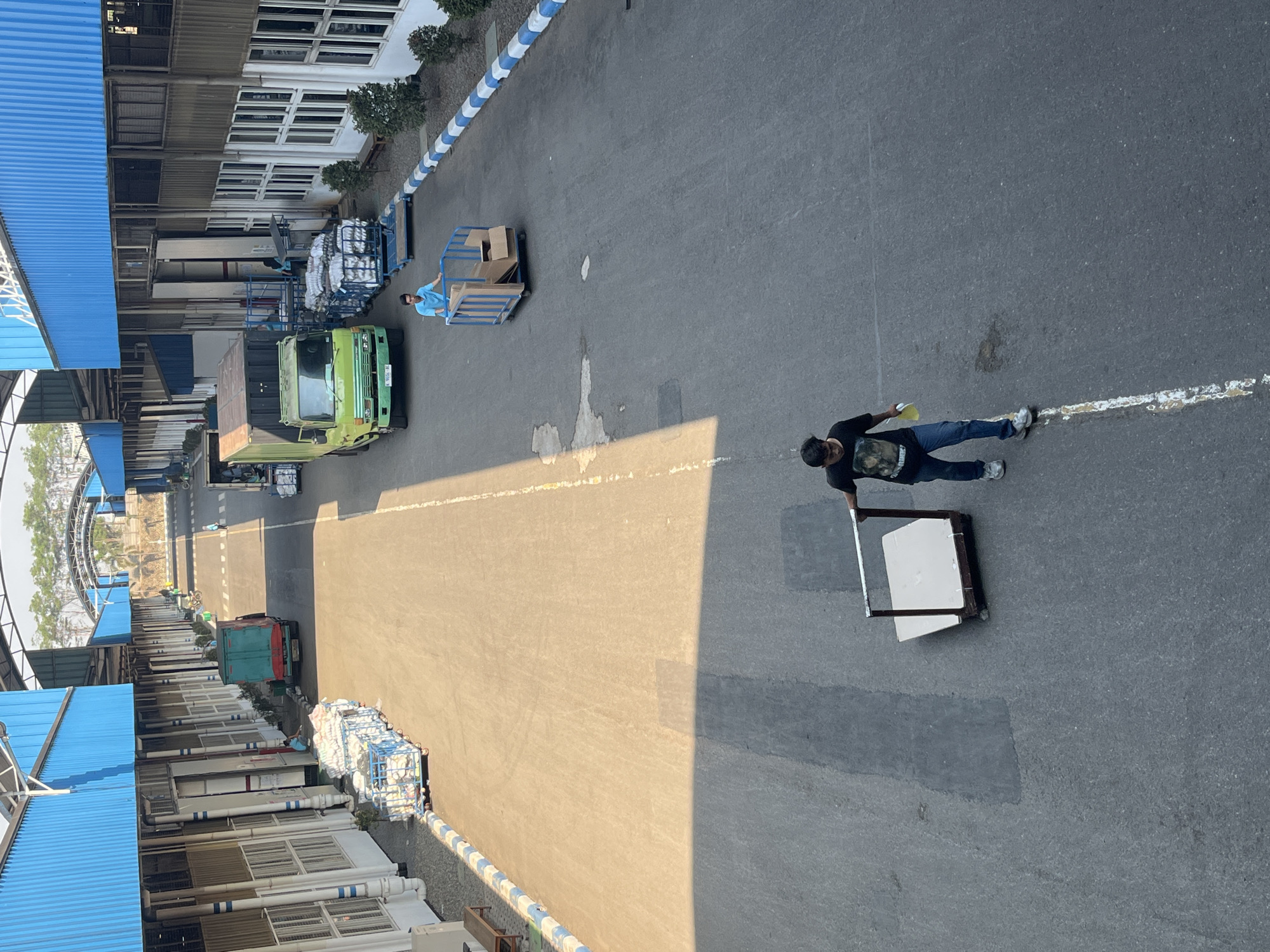 The image shows an outdoor industrial area, likely a logistics or manufacturing facility, in Pelang, Indonesia. In the foreground, a young man wearing a black t-shirt with a graphic print and blue jeans is walking away from the camera, pushing a large, flat-topped weighing scale. He is holding a yellow object in his left hand. Further up the asphalt road, another young man in a bright blue t-shirt is pushing a blue metal cart containing several cardboard boxes. Behind him, a green and black truck is parked. To the left of the road are long, single-story industrial buildings with blue corrugated metal roofs and beige walls. Along the edge of the road, a blue and white striped border delineates the walkway. In the background, a larger arched structure is visible, and beyond that, trees and a bright sky. The lighting suggests it is daytime, with strong shadows cast across the road.