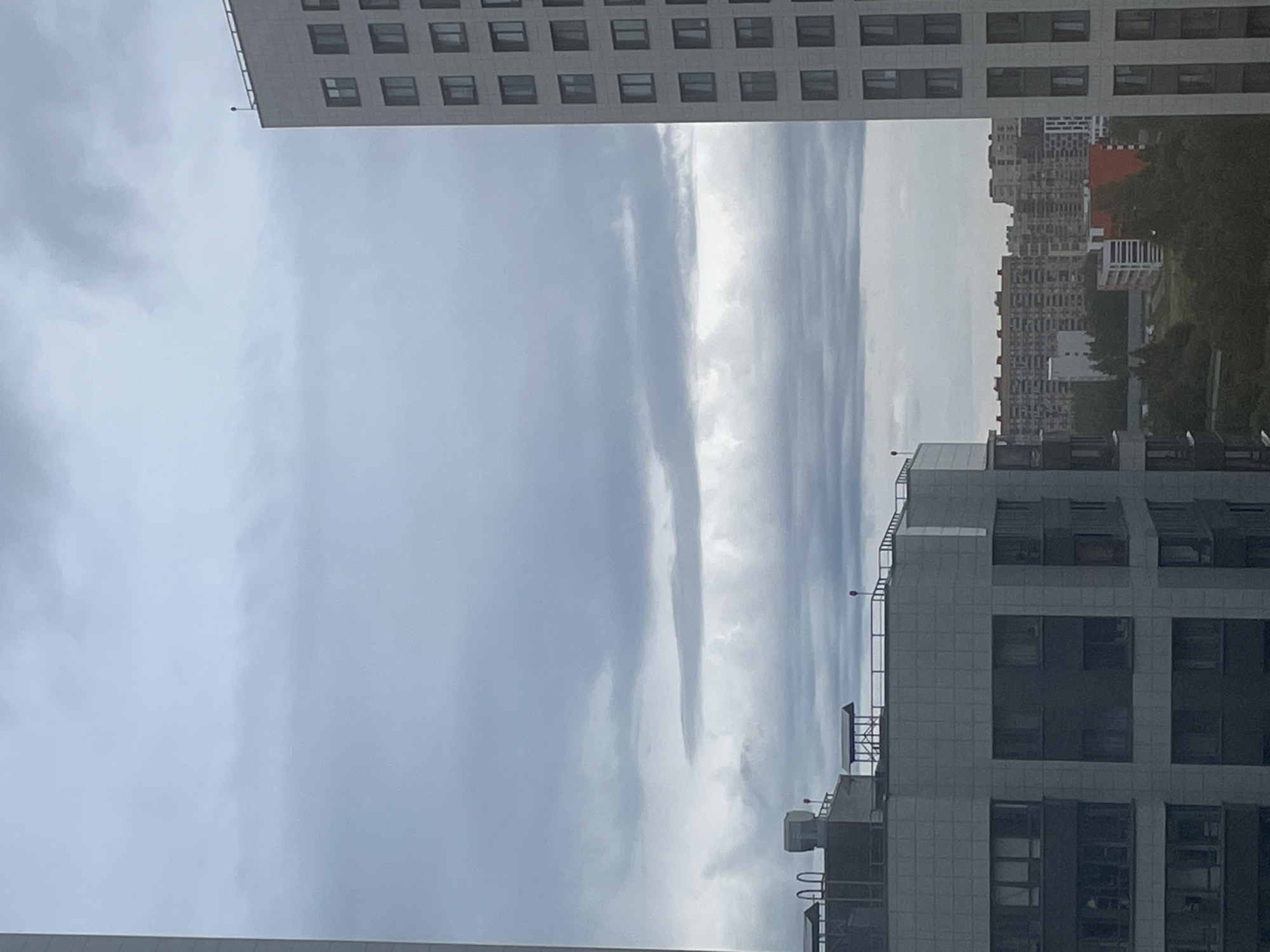The image shows a view of a city skyline under a cloudy sky, with several modern residential buildings. The foreground features parts of two tall, light-colored apartment buildings. The building on the right is a tall, rectangular structure with numerous windows. The building on the left is partially visible and appears to be connected to a lower, multi-story structure with a flat roof. In the middle ground, a complex of older, densely packed apartment buildings with a consistent architectural style is visible, interspersed with trees and some newer, more modern buildings. The background consists of a layered cloud formation across the sky. The scene appears to be a residential area in the city of Sinyavino, Russia. There are no people or animals visible in the image.