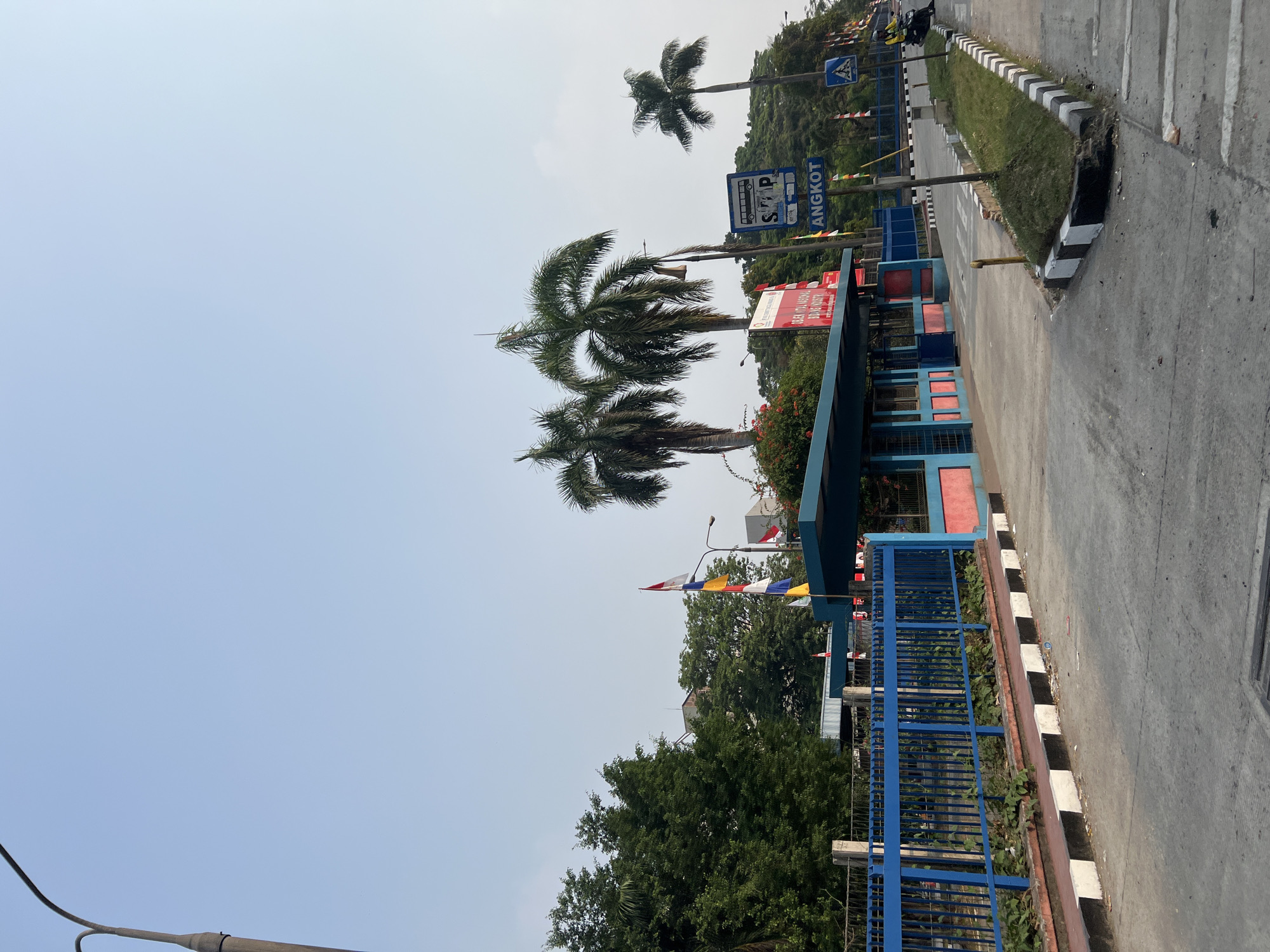 A low-angle, full shot captures a bright, sunny day at an outdoor facility entrance in Citeureup, Indonesia. The scene is dominated by a clear blue sky and a paved road. In the foreground, the road curves gently to the right, with a black and white striped curb separating it from a grassy area. Several traffic signs are visible, including a blue sign with a bus icon and the word "ANGKOT," indicating a public transportation stop. To the left, a blue metal fence encloses the property, behind which are trees and some buildings. A small, covered structure with blue and pink accents appears to be a guardhouse or entrance booth. Two palm trees stand prominently near the entrance. In the distance, a person on a yellow motorcycle is seen on the road. Several flags are displayed on poles.