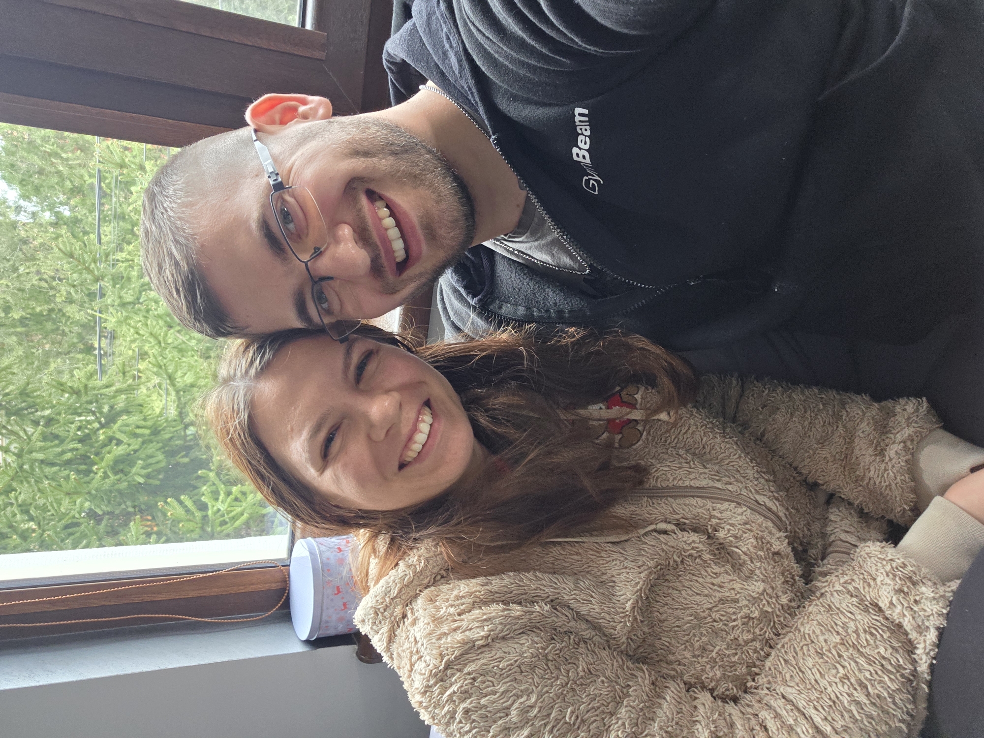 Two people are posing for a photo indoors. A man with short grey hair, glasses, a beard, and wearing a black hoodie with the word "GymBeam" on the sleeve is positioned above a woman. The woman has long brown hair and wears a furry, tan-colored jacket. Both individuals are smiling. Behind them, there is a window showing green trees. The location is likely in Yagodovo, Bulgaria, based on the prompt. A decorative box sits on the windowsill.