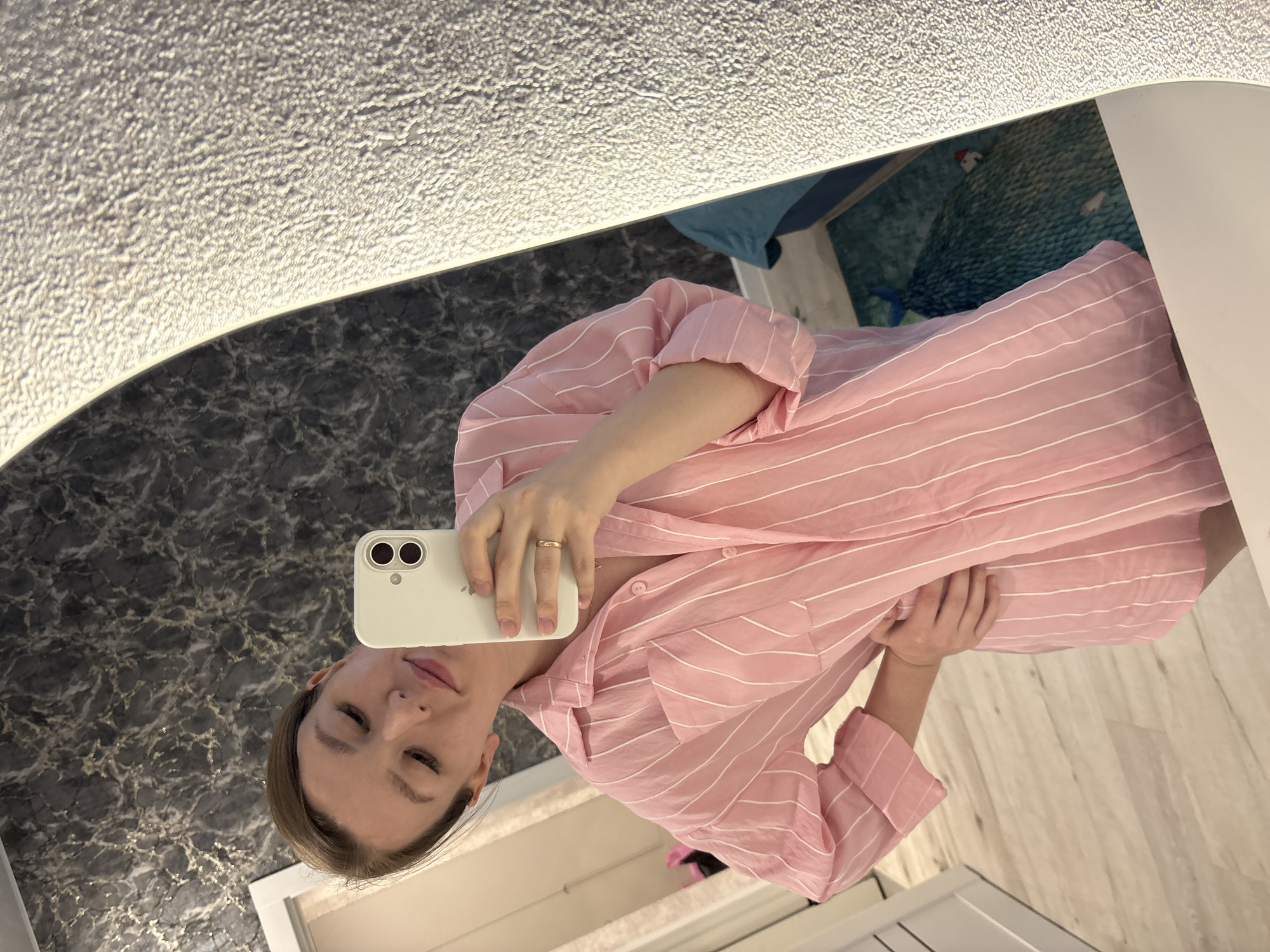 A young woman is taking a selfie in a bathroom mirror. She is wearing a pink and white striped shirt. The mirror has a light around it. There is a marble countertop below the mirror. A patterned rug is visible in the background. A white cabinet is partially visible in the foreground. The location is likely a private residence, and no evidence suggests it is in Izhevsk, Russia.