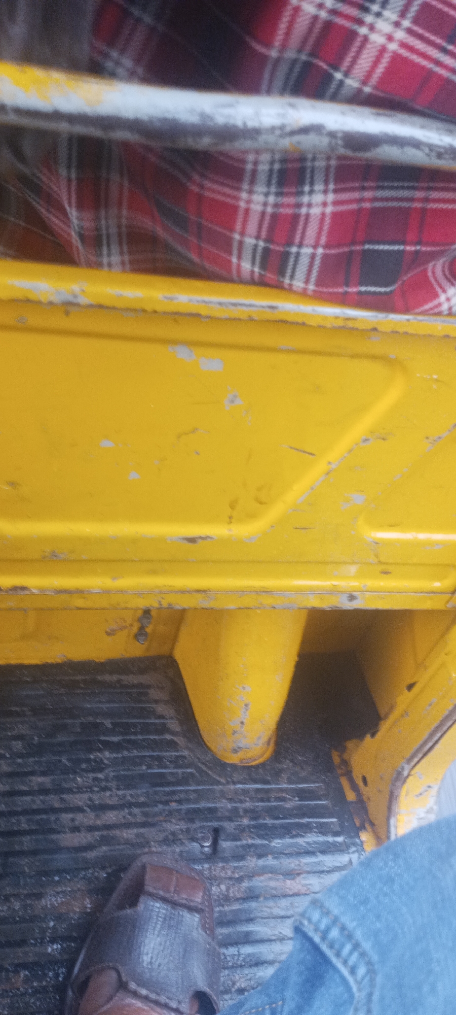 The image shows the interior of a vehicle, possibly an auto rickshaw or similar small transport, in Umuahia, Nigeria. The vehicle has yellow painted metal surfaces with areas of chipped paint. The floor is made of grooved metal. A person's foot wearing a brown sandal and clad in blue denim is visible. Above the yellow panel, a red and black plaid patterned fabric is present. The scene appears to be taken from inside the vehicle looking down at the footwell.