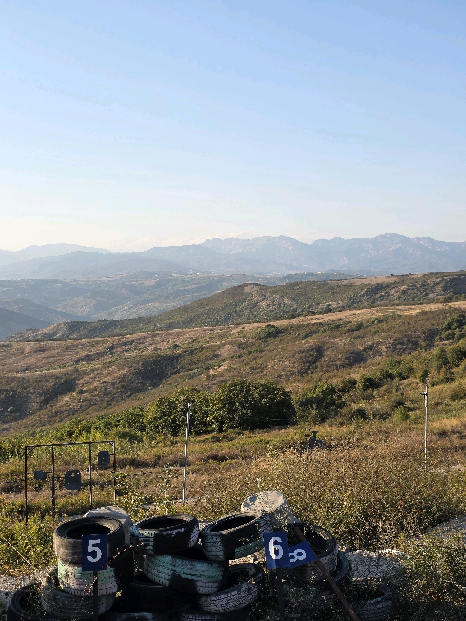 The image depicts a shooting range located in the mountainous terrain outside the city of Muğanlı, Azerbaijan. In the foreground, a pile of used tires serves as a barrier, with two blue signs displaying white numbers "5" and "6" attached to them. Behind the tires, a metal frame holds several target plates. The background features rolling hills covered in dry grass and scattered green trees, leading to distant, hazy mountains under a clear, light blue sky. The scene suggests an outdoor recreational or training facility.