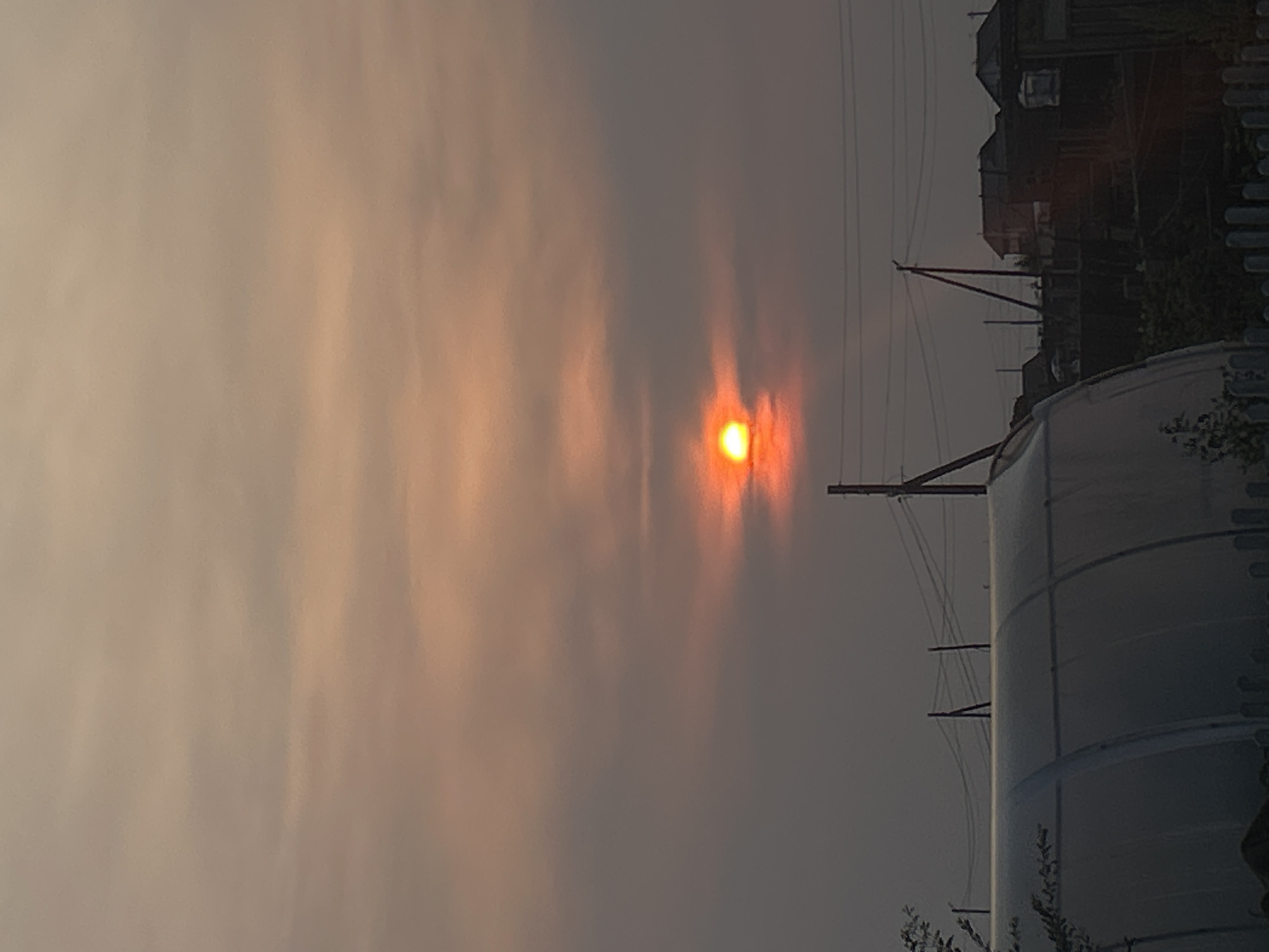 The image depicts a rural landscape at sunset, with the sun, a bright orange orb, partially obscured by hazy clouds. The sky is a gradient of pale orange and grey. In the foreground, a large, arched structure, possibly a greenhouse, dominates the lower left portion of the frame. Behind it, silhouetted against the dimming sky, are several small houses and utility poles with electrical wires. The overall impression is one of a quiet, possibly agricultural, setting in a town like Appany, Russia. No people are visible in the image.