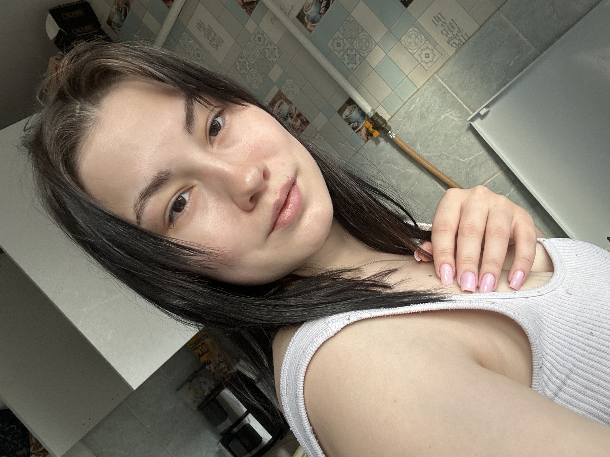 A close-up, angled selfie depicts a young woman with dark hair and fair skin. She wears a white ribbed tank top and displays pink nail polish. The background includes a wall with decorative tiles and a white refrigerator or cabinet. A pipe runs along the wall. The location is possibly a kitchen or bathroom in Chaikovsky, Russia. No specific interactions or actions are evident in the image.