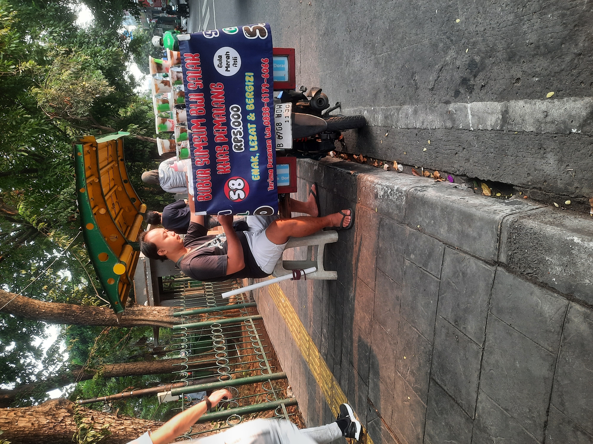 A medium shot captures a street vendor preparing to serve a customer from a mobile food cart on a city sidewalk. The vendor, a man with short dark hair, sits on a small plastic stool, wearing a dark t-shirt and grey shorts. He is facing left, holding a white plastic cup with a straw and looking down at his hands. The food cart, mounted on a motorcycle, is parked to his right. A blue banner on the cart advertises "BUBUR SUMSUM BIJI SALAK KHAS PEMALANG" and lists the price as "RP5.000". Behind the vendor, two women are visible near the cart, one wearing a grey hoodie and the other a dark shirt. The sidewalk is paved with dark grey tiles and has a yellow tactile paving strip. The background features trees with lush green foliage and a metal fence. The scene appears to be set in North Jakarta, Indonesia, during the day, with natural light casting shadows.