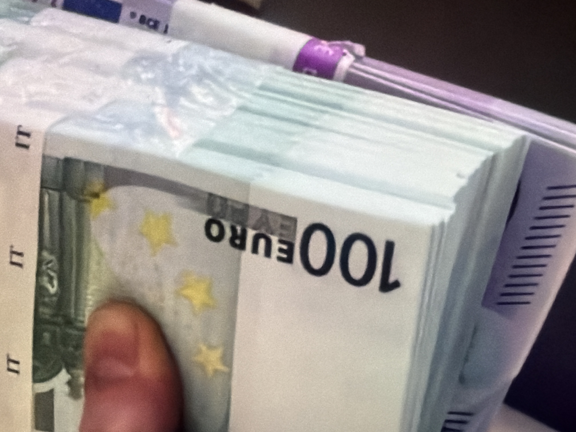 The image shows a close-up view of a stack of 100 Euro banknotes. The banknotes are partially wrapped in plastic. In the foreground, a human thumb is visible, lightly pressing on the banknotes. Some yellow stars are visible on the banknotes, which are a common design element on Euro currency. The "100 EURO" denomination is clearly printed on the banknotes. A purple band with some writing is visible on the right side of the stack. The background is dark and out of focus. The image's metadata or context does not provide any information about its location in Bukuru, Nigeria.