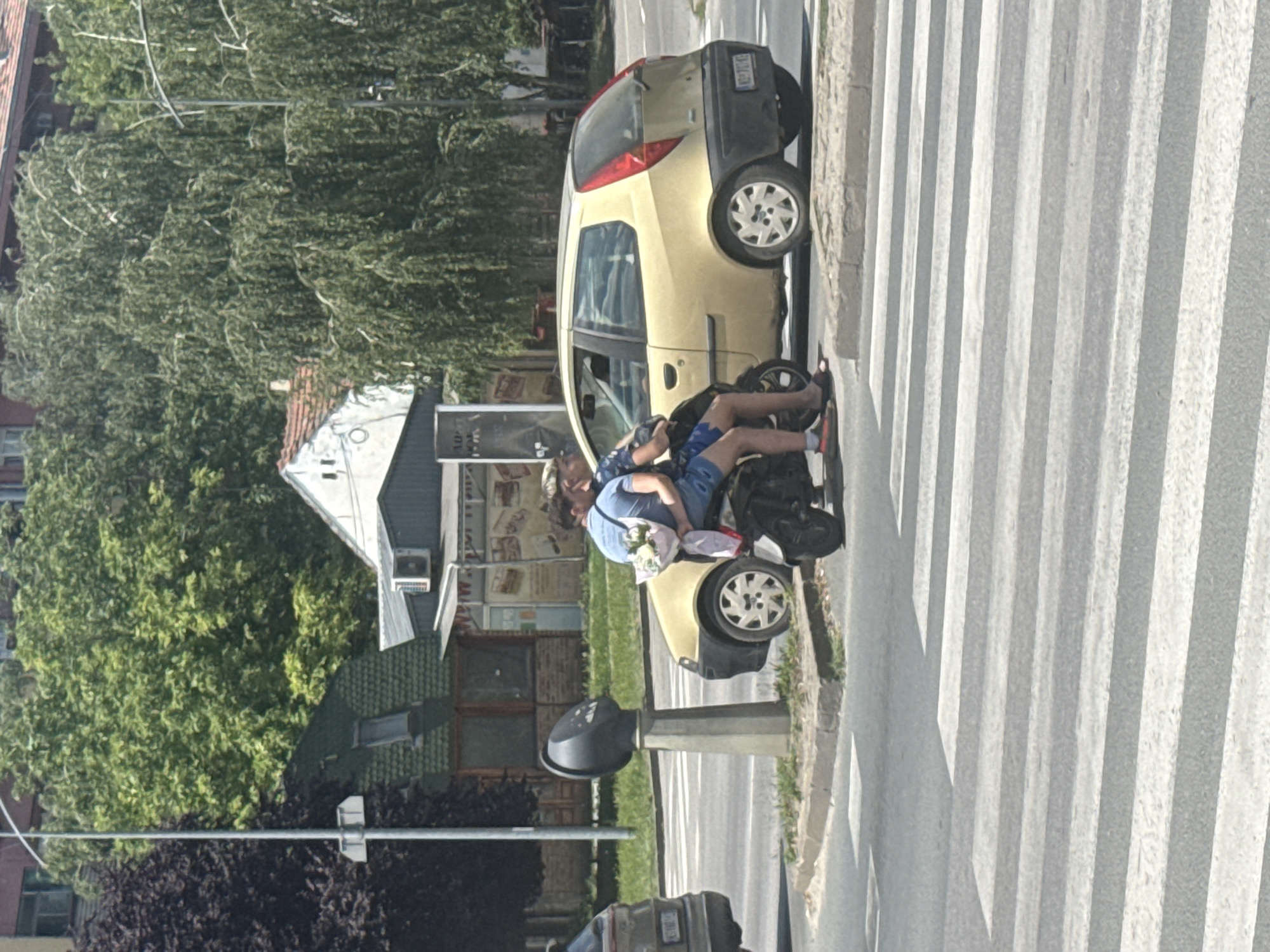 Two individuals, one driving a scooter and the other as a passenger, are stopped at a pedestrian crossing in Novi Sad, Serbia. The driver, wearing a blue t-shirt and dark shorts, is positioned in front. The passenger, wearing a patterned shirt and blue shorts, is behind the driver, holding a bouquet of white flowers wrapped in pink paper. They are seated on a black scooter. To their right is a yellow hatchback car. In the background, there is a payphone on a concrete pedestal. Beyond the payphone and car, there is a building with a green tiled roof and a white facade, and further in the distance, dense green trees and foliage. The sky is clear and appears to be daytime.