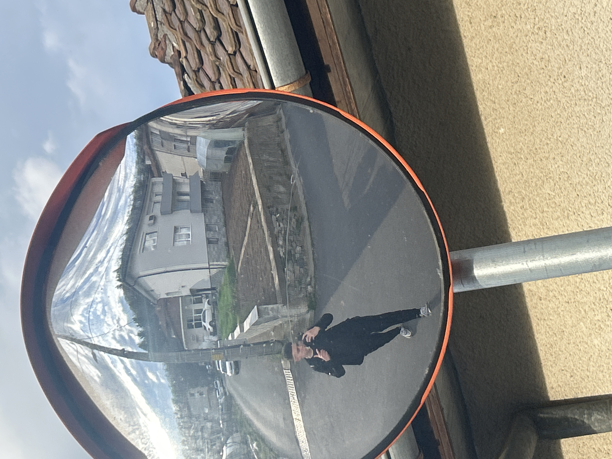 The image depicts a convex traffic mirror with an orange frame mounted on a metal pole near a building with a tiled roof. The mirror reflects a street scene, including buildings, cars, and a person dressed in dark clothing who is possibly taking a picture of themselves. The buildings appear to be located in a residential area. Based on the location context, it can be assumed that the image was captured in Zlatograd, Bulgaria.