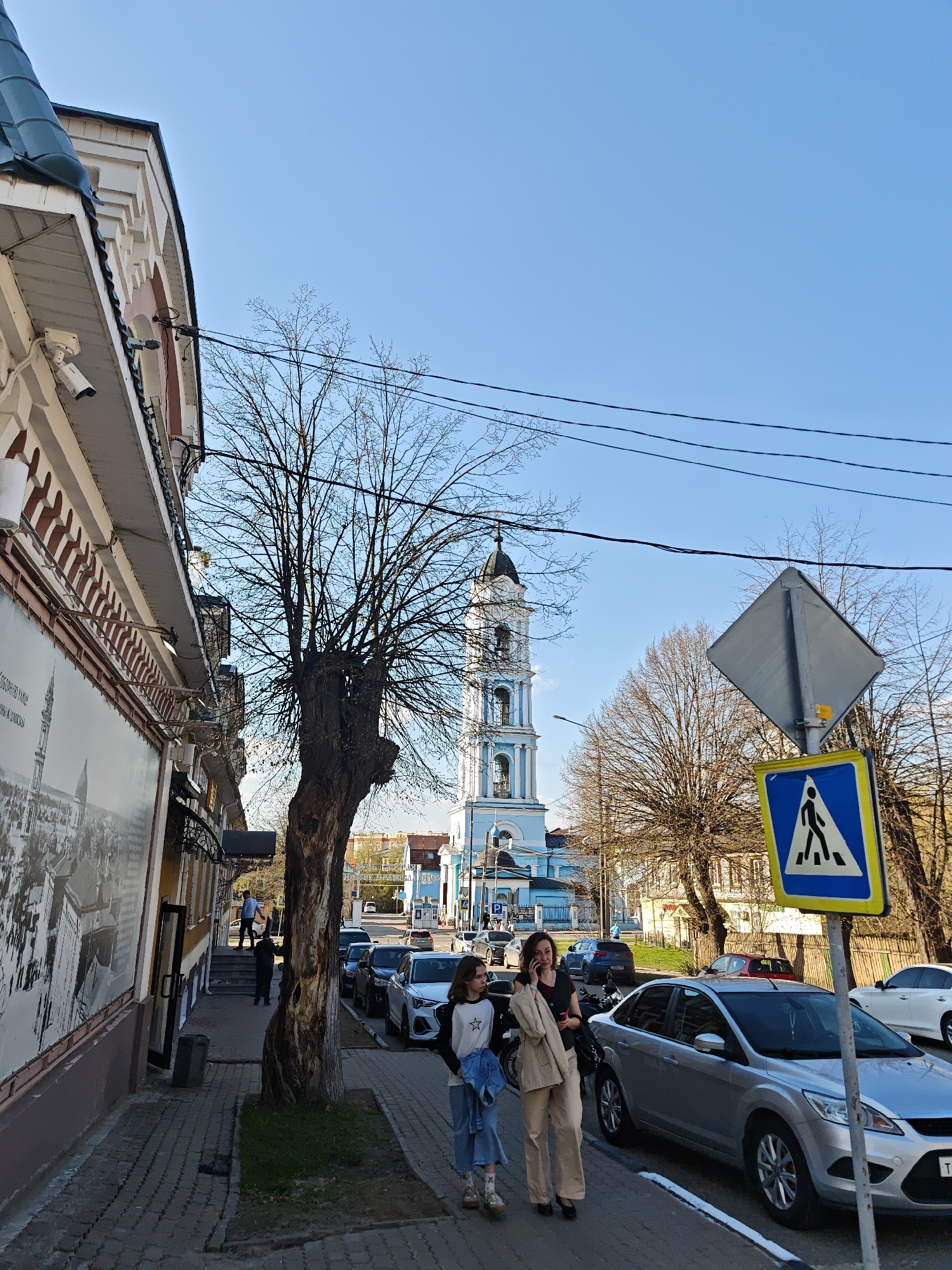 The image shows a street scene in Noginsk, Russia. In the background is a blue and white bell tower of the Epiphany Cathedral. A mother and daughter are walking along a sidewalk next to parked cars. The sidewalk runs along a building with a large black and white photograph on its side. A crosswalk sign is visible on the right side of the street. A bare tree is present between the sidewalk and the street. People are entering a building with steps. The sky is blue.
