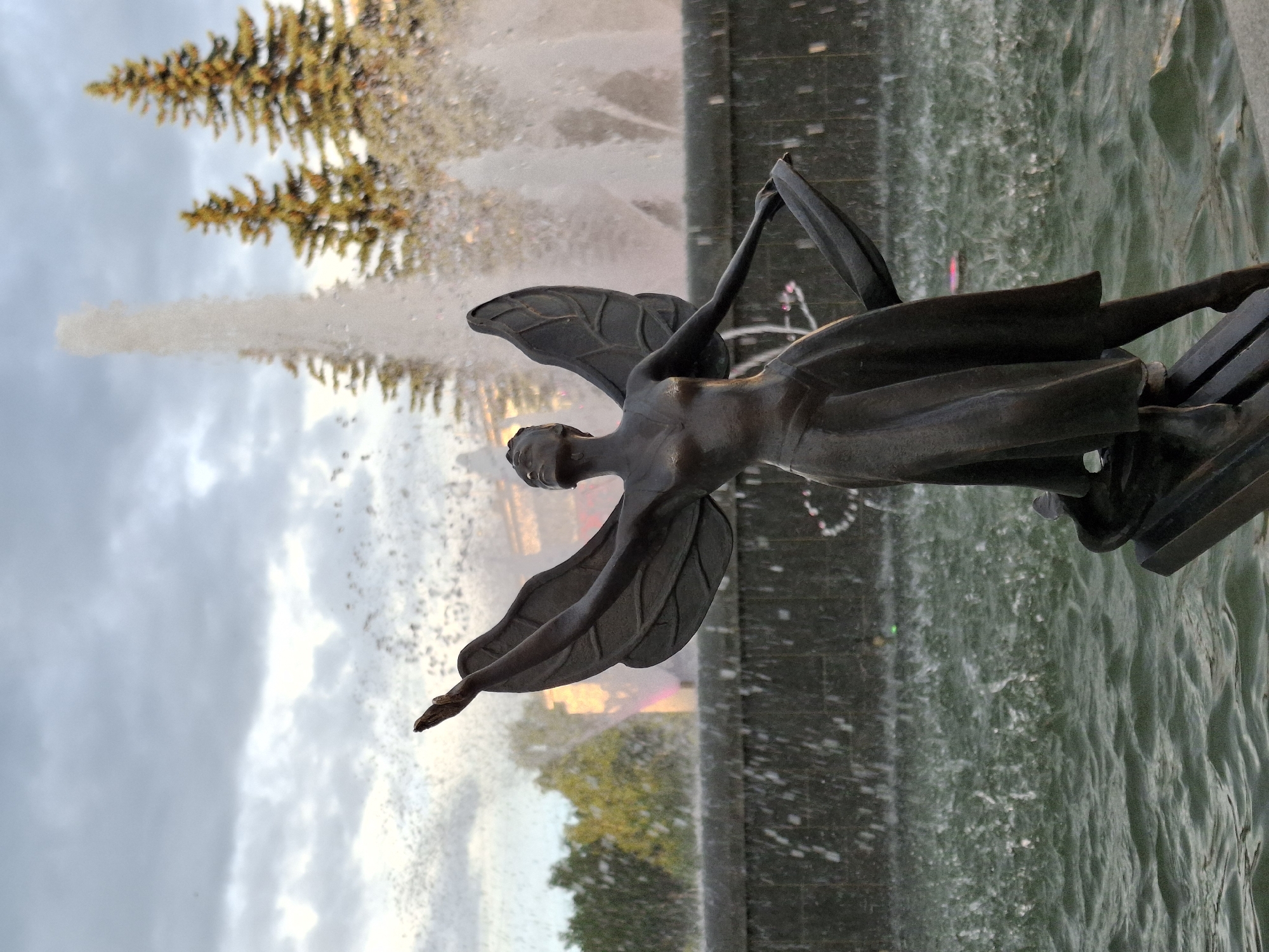 This media file shows a statue of an angel in a fountain. The angel is made of dark metal and has large wings. It is positioned in the center of the frame, with its arms raised. Water from the fountain is spraying around the statue, creating a misty effect. In the background, there are trees and a cloudy sky. The overall impression is one of grandeur and movement. The lighting suggests it is either dawn or dusk. The statue appears to be located in a public space, possibly a park or a square, given the presence of the fountain and trees. The water in the fountain is green, possibly due to algae or lighting. The sky is overcast with some patches of light breaking through, indicating a mix of sun and cloud, typical of temperate weather. There is no visible text in the image.