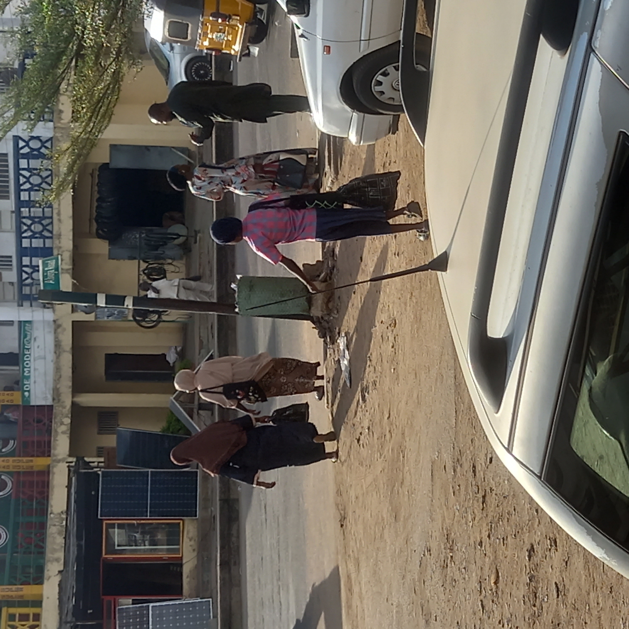 The image depicts a street scene in Dadi Riba, Nigeria. A man in a pink and black shirt and blue pants stands near a green trash bin, using a long stick to interact with the contents of the bin. Several other people are walking nearby, including a woman in a brown hijab and black dress. In the background, there is a building with solar panels displayed outside. A white car is parked on the street.