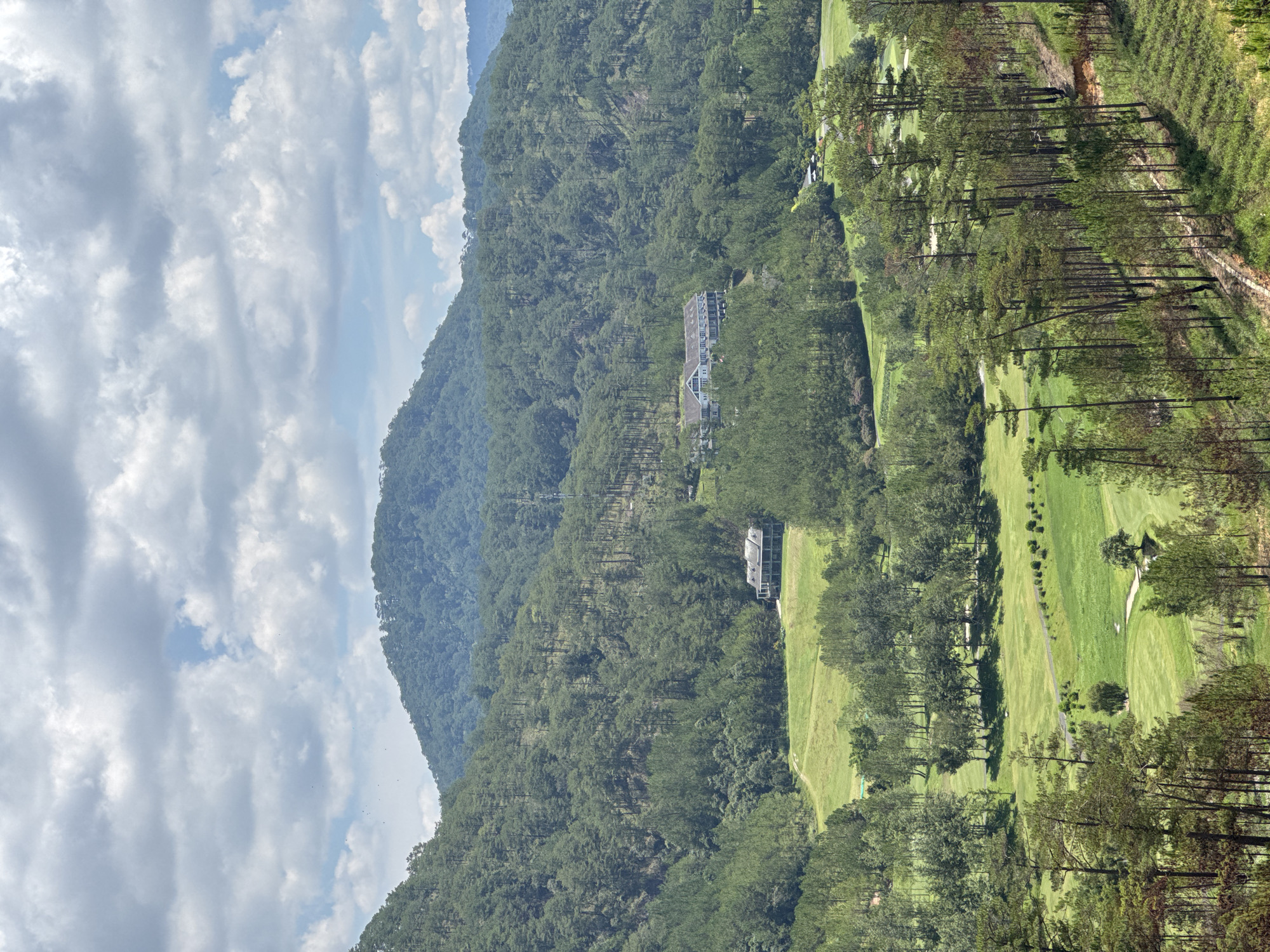 A landscape in Hiep Thanh, Vietnam, shows a green golf course surrounded by lush, green trees. The course is partially obscured by these trees. In the distance, a green mountain is visible, with dense trees covering its surface. A building sits nestled among the trees on the mountain. To the left, a cloudy sky with patches of blue is seen. The image is shot from an elevated perspective, providing a wide view of the terrain.