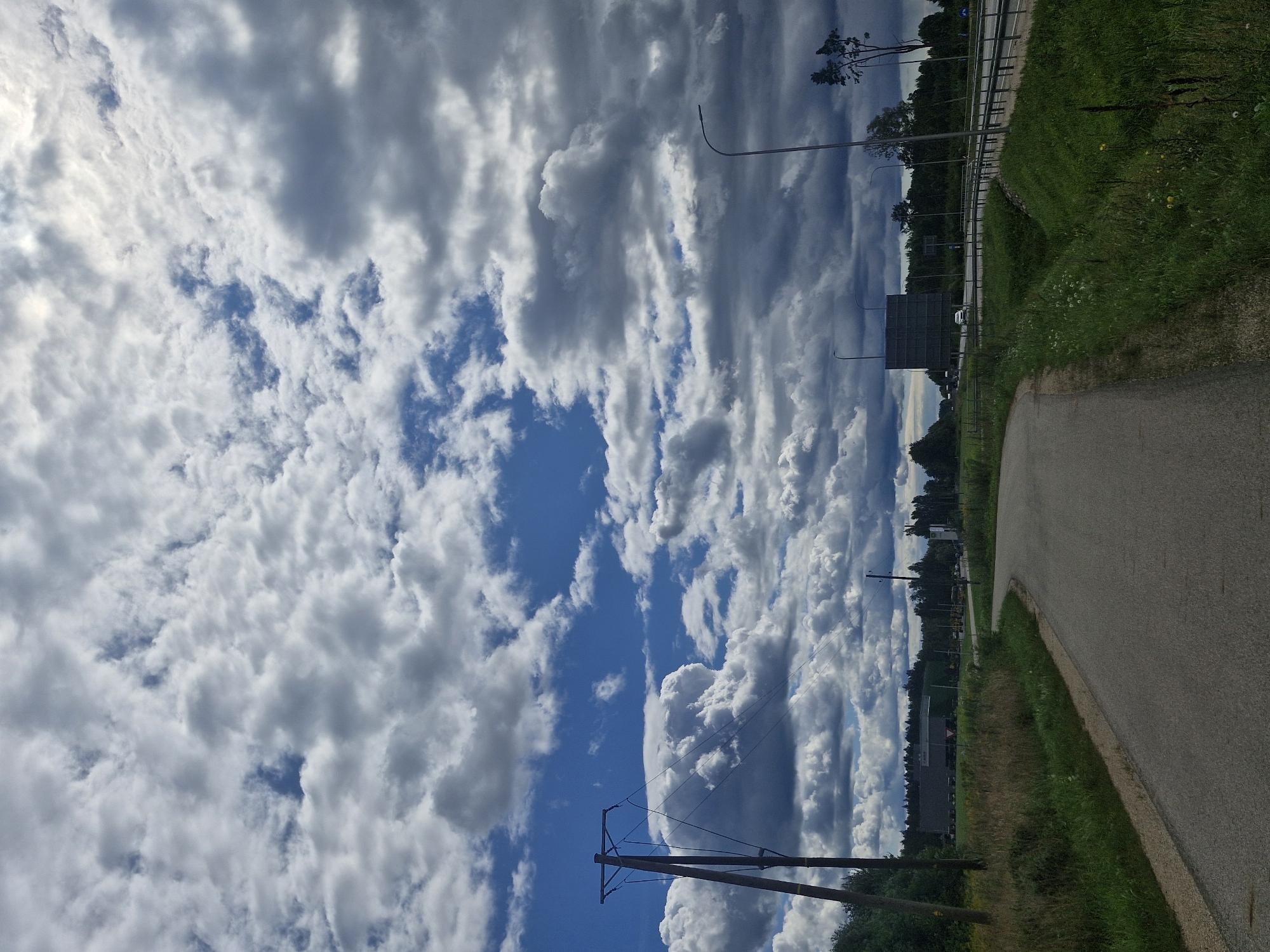 The image depicts a rural landscape on a cloudy day. A grey asphalt road curves from the foreground towards the left. On the left side of the road, a tall wooden utility pole with wires is visible, set against a background of green grass and trees. Further in the distance, a large, dark building with a flat roof is situated among a line of trees. On the right side of the road, a guardrail separates the road from a grassy embankment. A modern streetlamp stands on the right side, and a dark, rectangular billboard structure is visible in the distance beyond the guardrail, framed by trees. The sky is filled with dramatic, dark grey and white cumulus clouds, with patches of blue sky visible in between.