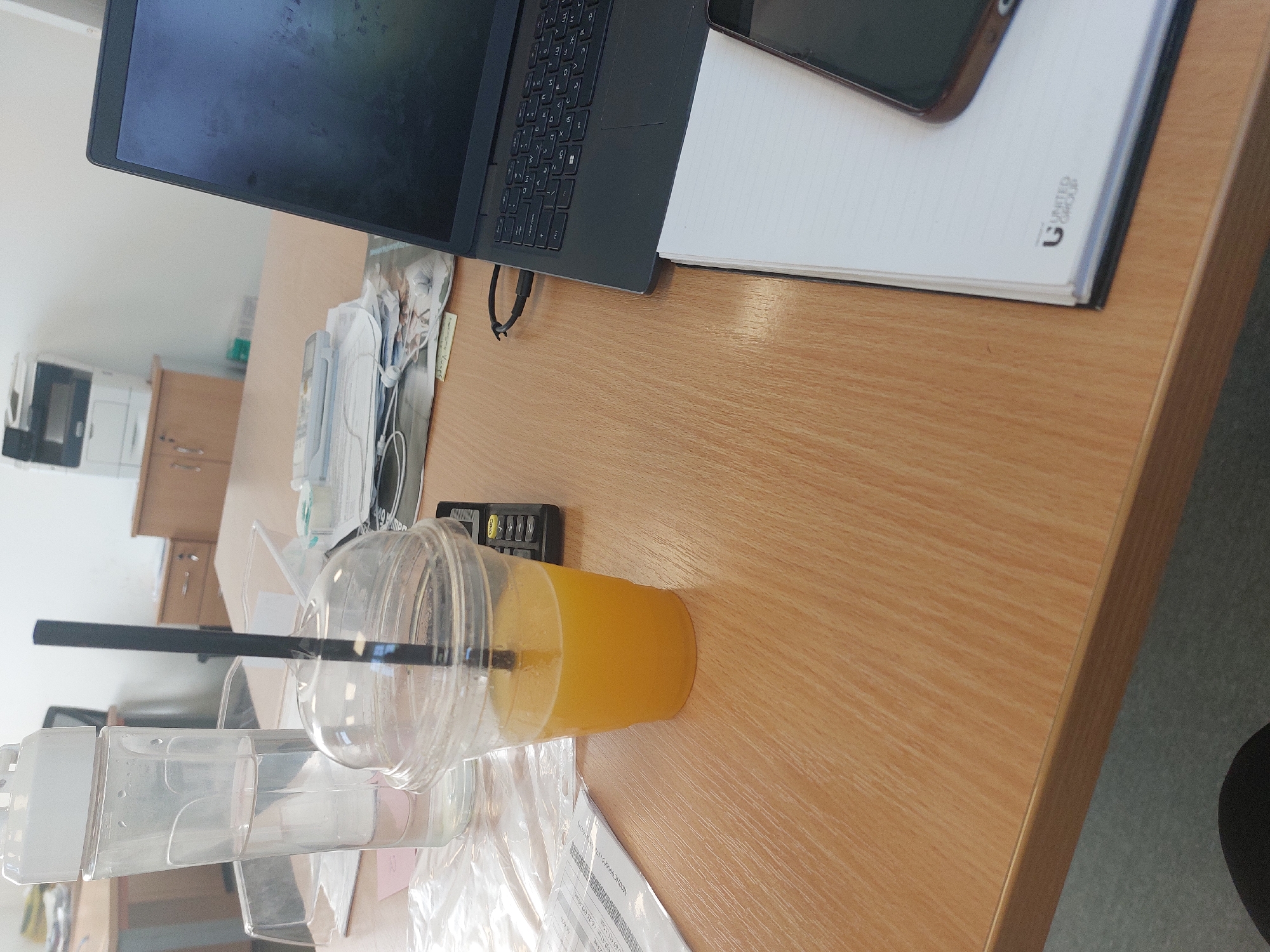 The image shows a desk with a laptop, a notebook, a phone, a cup of orange juice with a straw, and a water bottle. Other items are scattered around the desk, including what appears to be packaging and some other small items. The desk is in an office setting. While it is impossible to pinpoint the exact location in Burgas, Bulgaria from the image alone, the presence of typical office equipment suggests the image may be from a professional workspace.