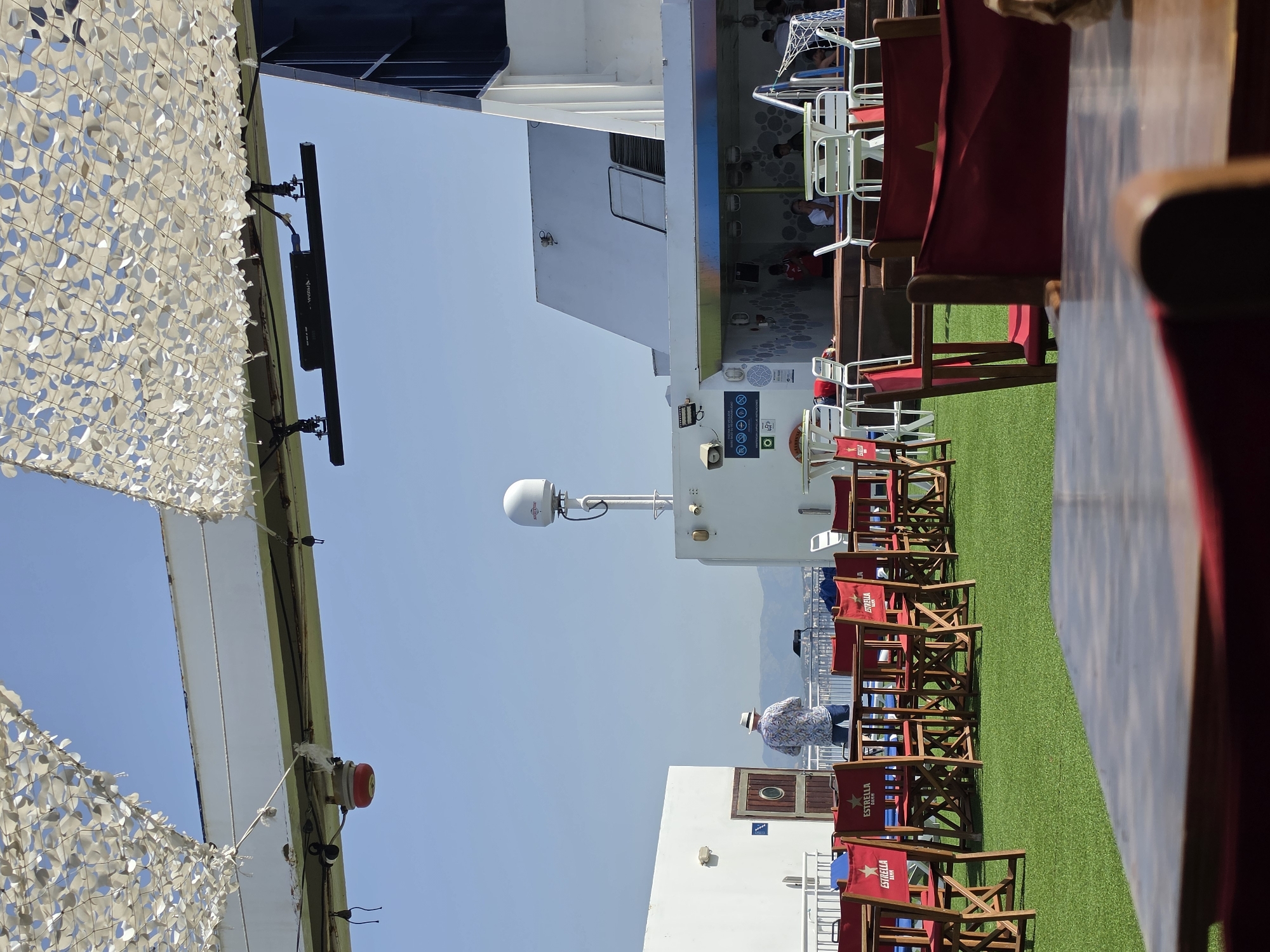 The image depicts an outdoor deck area of a ferry or cruise ship, under a camouflage netting canopy, on a bright, sunny day. The deck is covered with artificial turf and furnished with numerous wooden tables and red chairs, many of which bear the "ESTRELLA DAMM" logo. In the background, a white ferry structure rises, with a satellite dish visible atop it. A man in a floral shirt and hat stands on the deck, facing away from the camera, appearing to interact with an object on a table. Further in the background, other passengers are seated at tables in a shaded area. The distant horizon shows a hazy outline of hills or mountains. The location is implied to be the city of Magaluf, Spain, suggested by the presence of "ESTRELLA DAMM," a Spanish beer brand popular in tourist areas.