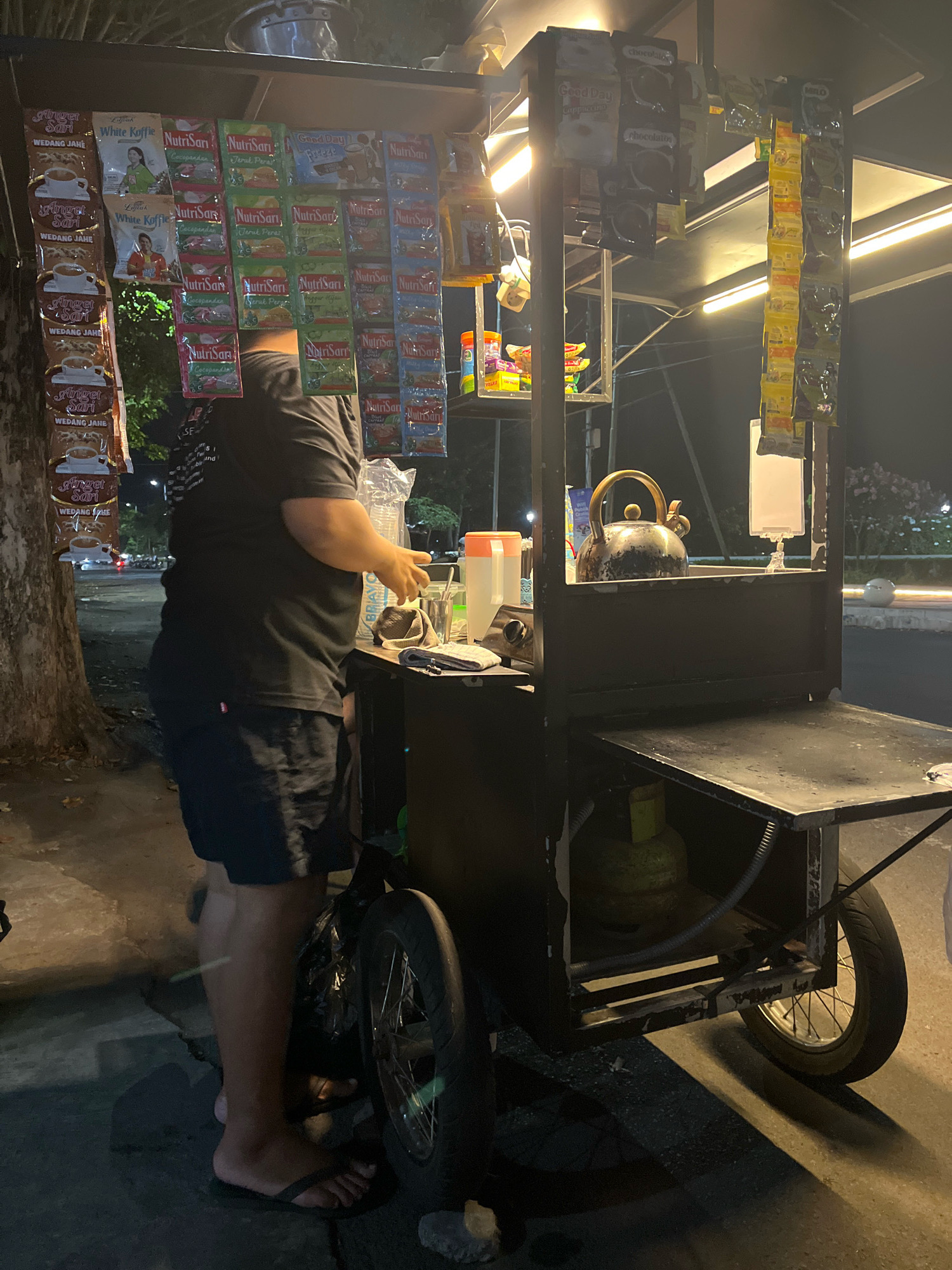 This image depicts an outdoor scene at night, likely on a street in Lumajang, Indonesia, illuminated by artificial lighting from a food cart and overhead streetlights. The focal point is a mobile food cart, a common sight in Indonesian cities, selling a variety of instant beverage packets, including "NutriSari" and "White Koffie" brands, along with other packaged goods hanging from its structure.
A person, wearing a black t-shirt and dark shorts, stands near the cart, facing away from the camera. They appear to be interacting with items on the cart, possibly preparing a drink. The cart itself is constructed from dark metal and features a serving counter, a kettle, and a gas cylinder stored underneath. The ground is paved with asphalt, and a tree trunk is visible to the left of the cart. In the background, blurred lights suggest ongoing street traffic. The overall atmosphere is that of a typical nighttime street scene in a populated Indonesian urban area.