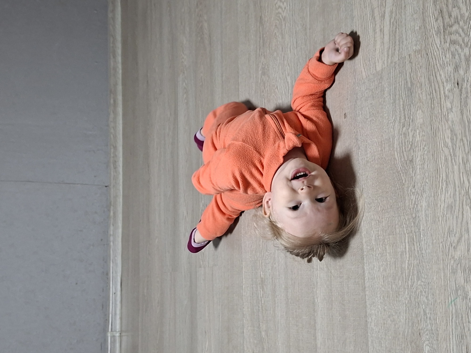 A young child wearing an orange fleece pajama set and purple socks is lying on their back on a light wood-grain floor. The child is smiling and looking up, with one arm raised in a fist. The scene appears to be indoors, possibly in a home or apartment. A portion of a gray wall is visible on the left side of the frame. The specific location is Unknown, Unknown.