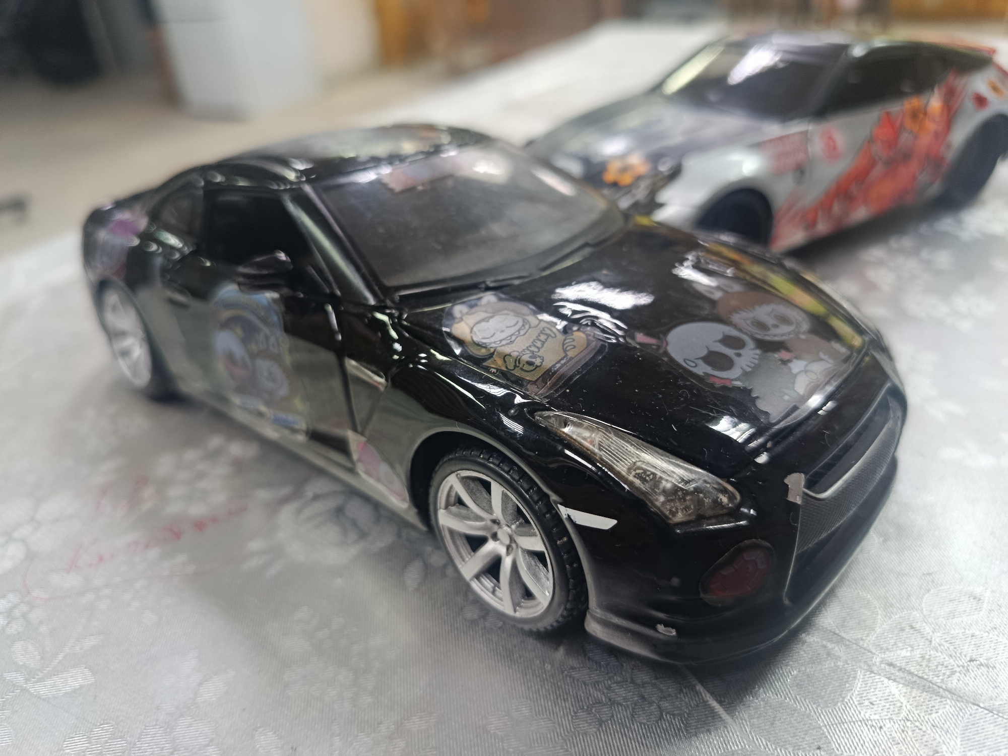 Two toy cars are placed on a textured surface. The car in the foreground is black and appears to be a Nissan GT-R. It has decorative stickers on its hood, including skulls and cartoon characters. The car in the background is silver and has red decals. The background is blurred and appears to be indoors. There is no indication of Sertolovo, Russia in the image.