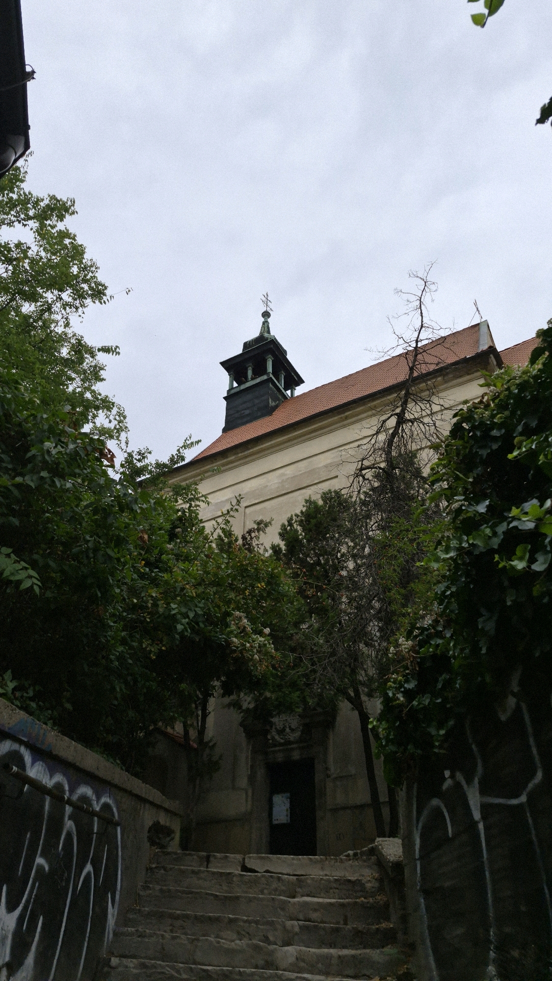 The image shows an outdoor view of a building in Bratislava, Slovakia, with a street leading up to it. The building appears to be a church or a historical edifice, with a distinct bell tower topped with a cross. The facade is light-colored, and the roof is tiled with red tiles. Lush green trees and foliage surround the structure, partially obscuring it. The foreground features a stone staircase with graffiti on a low wall to the left and a dark wall on the right, also bearing graffiti. The sky is overcast, suggesting a cloudy day. There are no people visible in the image.