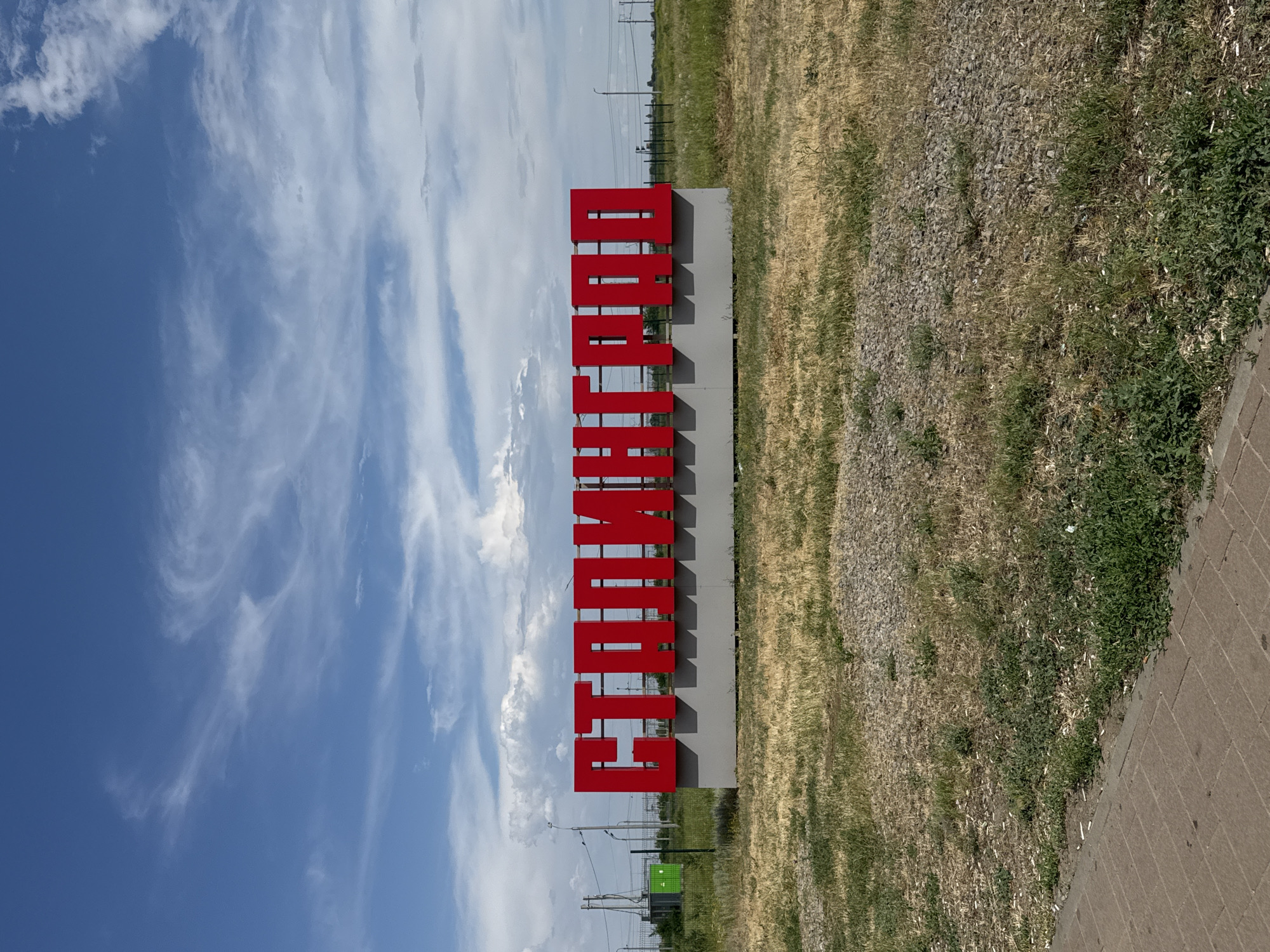 A large sign with red, three-dimensional letters spelling "СТАЛИНГРАД" (STALINGRAD) is centrally placed in a grassy, somewhat overgrown field under a partly cloudy blue sky. The letters are mounted on a grey, rectangular base. In the background, to the left of the sign, there is a green electrical transformer box. Further in the background, there are power lines and poles, as well as a fence. The ground in front of the sign is a mix of dry grass, gravel, and some patches of green weeds. To the left foreground, a brick-paved walkway is visible. The overall environment appears to be an outdoor, possibly semi-industrial or roadside location on the outskirts of Volgograd, Russia, given the presence of the city's name on the prominent sign. There are no people visible in the image.