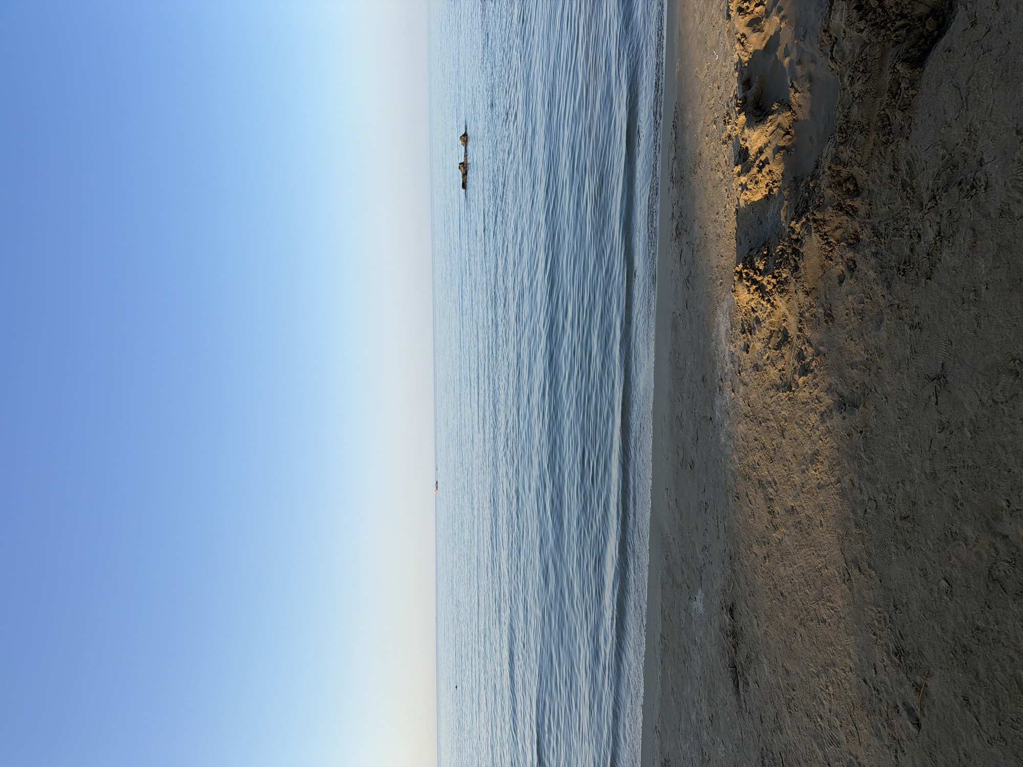 The image shows a wide, tranquil expanse of the sea and a sandy beach in Georgioupoli, Greece. The sky is a clear, pale blue, transitioning to a lighter hue near the horizon. The sea is calm with gentle ripples, reflecting the sky. In the distance, a few small boats are visible on the water. To the right, a cluster of dark rocks juts out from the water. The foreground is dominated by a sandy beach with scattered debris and mounds of sand, indicating it may be a well-used area for recreation. The lighting suggests it is either early morning or late afternoon, casting a warm glow on the sand. There are no people clearly visible in the image, but there are some faint footprints on the sand.