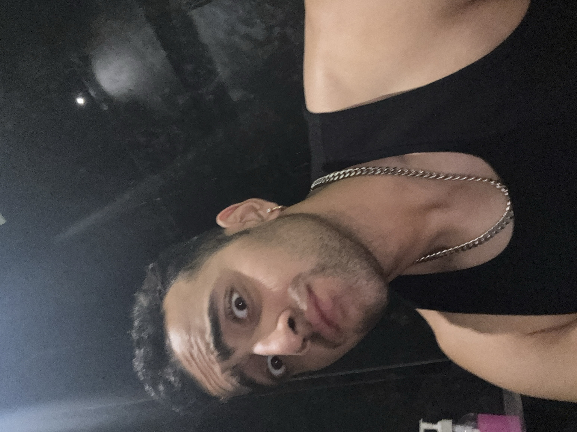 A selfie captures a man in a black tank top, wearing a silver chain necklace. His hair is dark and closely cropped. The background is dark and indistinct. The man appears to be indoors, with an unidentifiable white object and a pink item visible in the lower right corner. There are no location indicators to suggest the image was taken in Kolkata, India.