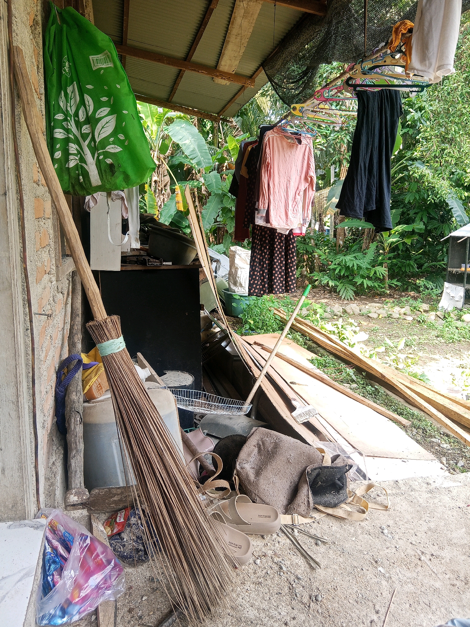The image depicts an outdoor storage area adjacent to a building. A large, green bag with a white tree graphic hangs on the left side, next to a rough brick wall. In the center, a tall broom with a wooden handle leans against the wall and a large gray container. Clothing items are visible hanging on a clothesline under a corrugated metal roof extension in the background, including a pink long-sleeved shirt and a black garment. A collection of wooden planks lies on the ground to the right of the broom. Several miscellaneous items are scattered on the ground, including a plastic bag filled with colorful items, what appears to be a small woven basket, and a pair of beige flip-flops. The ground is covered in a mix of dirt, sand, and small stones. Lush green vegetation is visible behind the storage area, suggesting a semi-rural or tropical environment. There are no people visible in the scene. The setting appears to be an informal outdoor storage space in Jinang Kampung Pansur, Indonesia, used for household items and cleaning tools.
