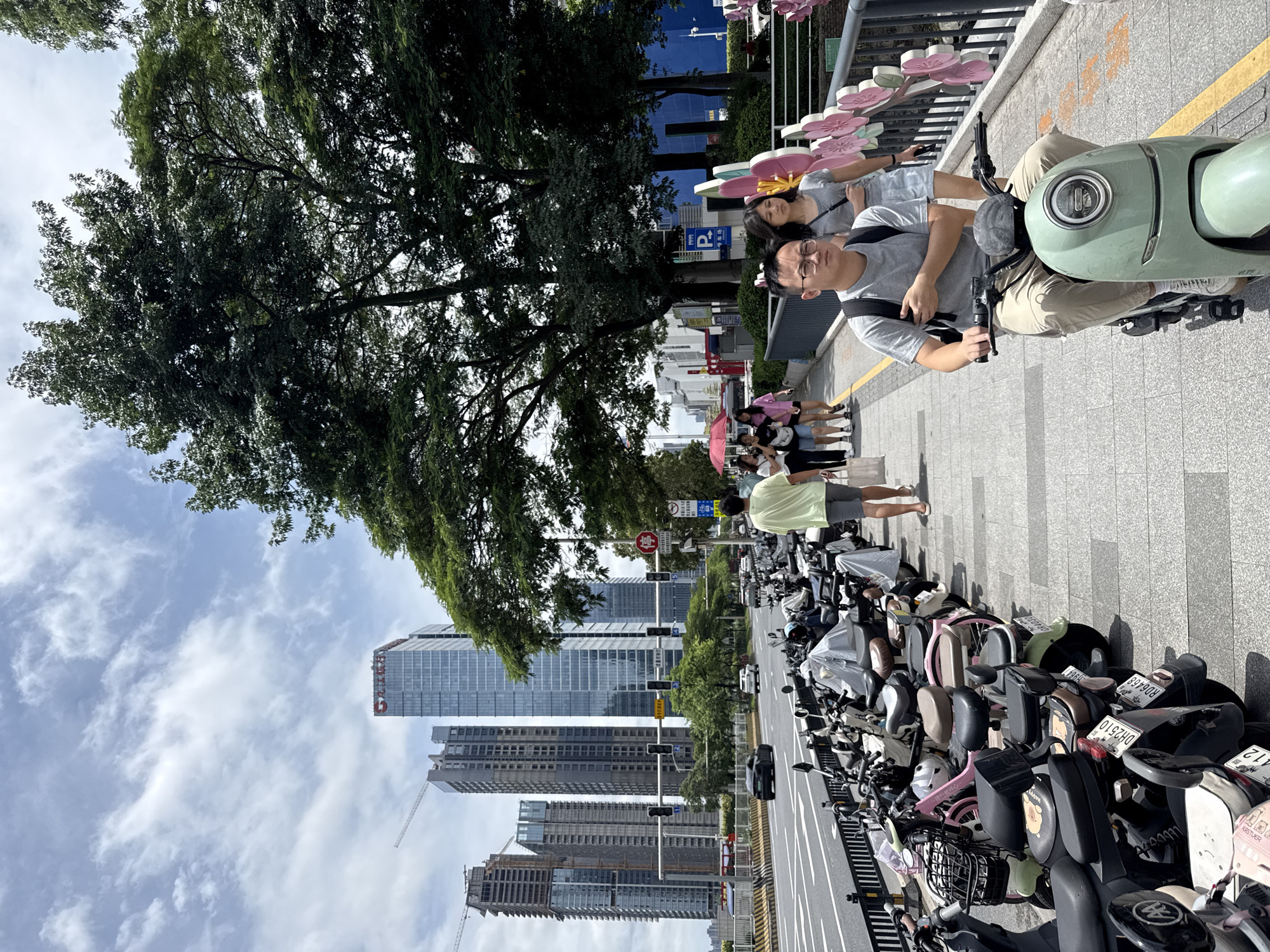 The image depicts a street scene in Tianhe District, China, on a cloudy day. A sidewalk runs alongside a busy road where several cars are visible. Parked along the sidewalk is a large row of electric scooters, some covered with tarps. Several people are walking on the sidewalk, including a man wearing glasses and a grey t-shirt riding a light green electric scooter, and a woman in a grey t-shirt and a light blue skirt walking behind him. Further down the sidewalk, a group of women are walking, one holding a pink umbrella. In the background, tall modern buildings and construction cranes are visible, indicating an urban environment. Lush green trees line the street and sidewalk, providing shade. A decorative pink floral barrier is visible on the right side of the sidewalk.