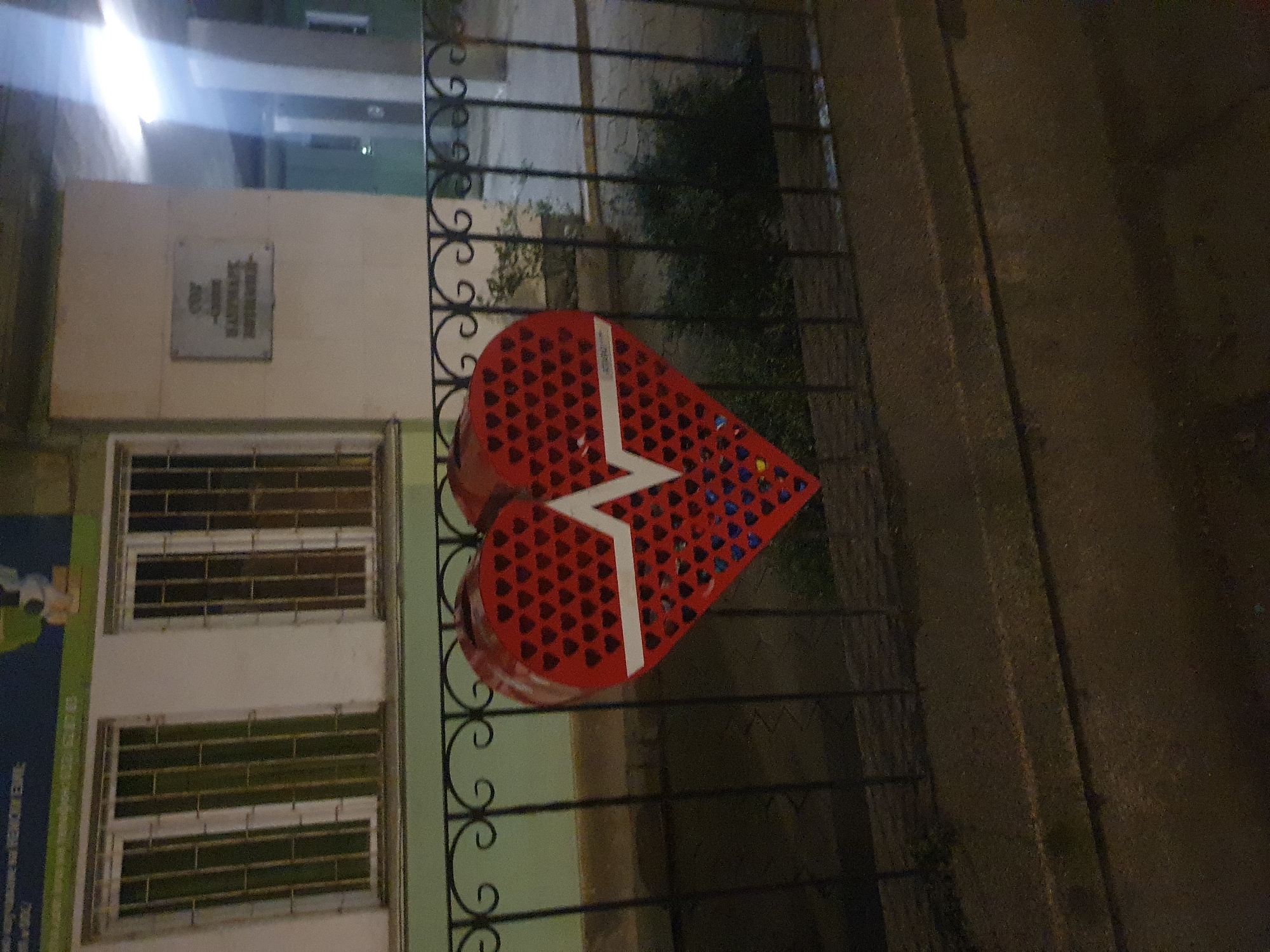A red heart-shaped art installation hangs on an iron fence in Plovdiv, Bulgaria. The heart is made of small, heart-shaped pieces and features a white lightning bolt design. The fence is black and ornate, with a building behind it. The building has white walls and barred windows. A plaque is visible on the wall above the windows. The lighting suggests it is nighttime. A green advertisement or sign is visible on the left side of the frame. The background is dark and indistinct, indicating an urban environment.