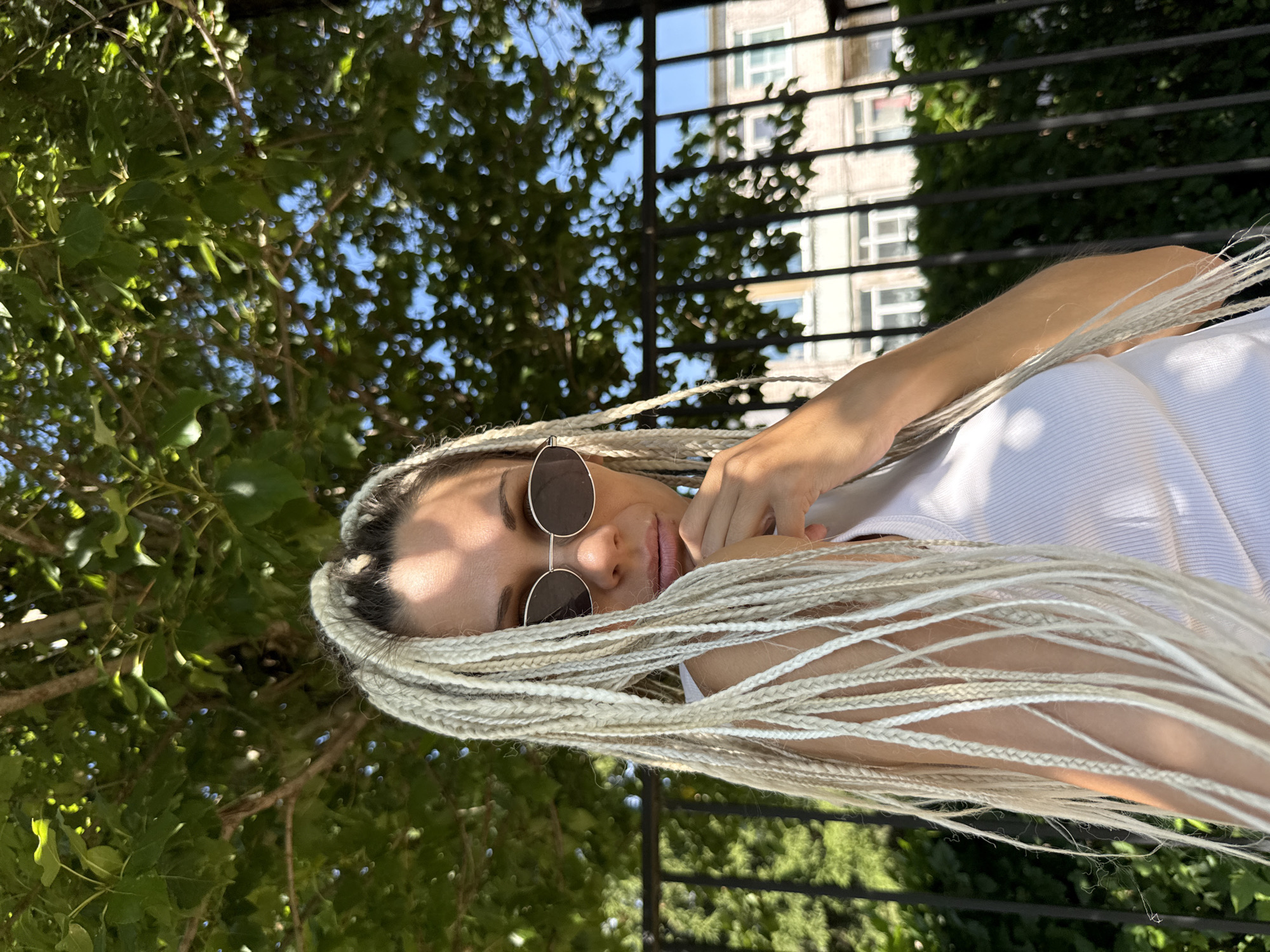 An outdoor, medium shot shows a woman from the chest up, facing the camera. She has light-colored braided hair that falls around her face and shoulders. She wears a white tank top and round, dark sunglasses. Her right hand is raised to her chin, with her fingers touching her lips. In the background, a black metal fence runs horizontally across the image. Behind the fence, there are trees with green leaves and a building with light-colored walls and windows. The sky is visible through the leaves and branches. The scene appears to be in a city park or garden.