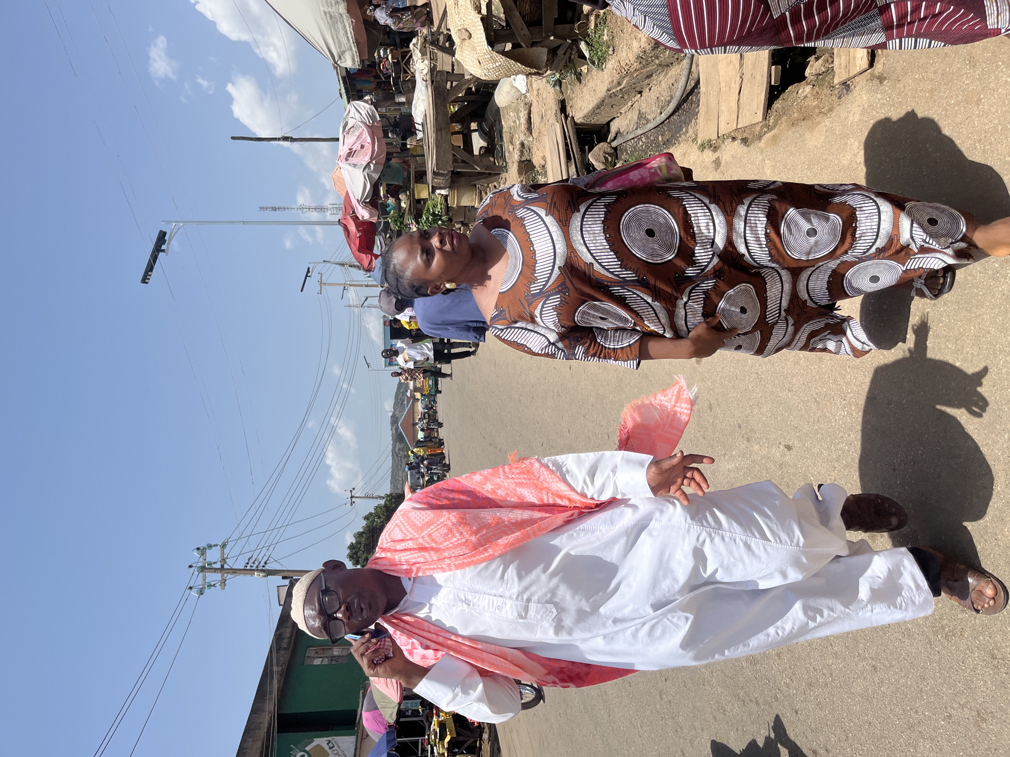 The image captures a street scene in Gwong, Nigeria, likely a market area due to the presence of stalls and vendors. In the foreground, a man wearing a white kaftan and a pink patterned scarf is walking and talking on a mobile phone. He has glasses and a white cap. Beside him, a woman in a brown and white patterned dress is walking. In the background, there are various market stalls with umbrellas, people walking, and motorcycles. The environment appears to be an outdoor urban setting with a paved road and buildings lining the sides. The sky is blue with scattered clouds.