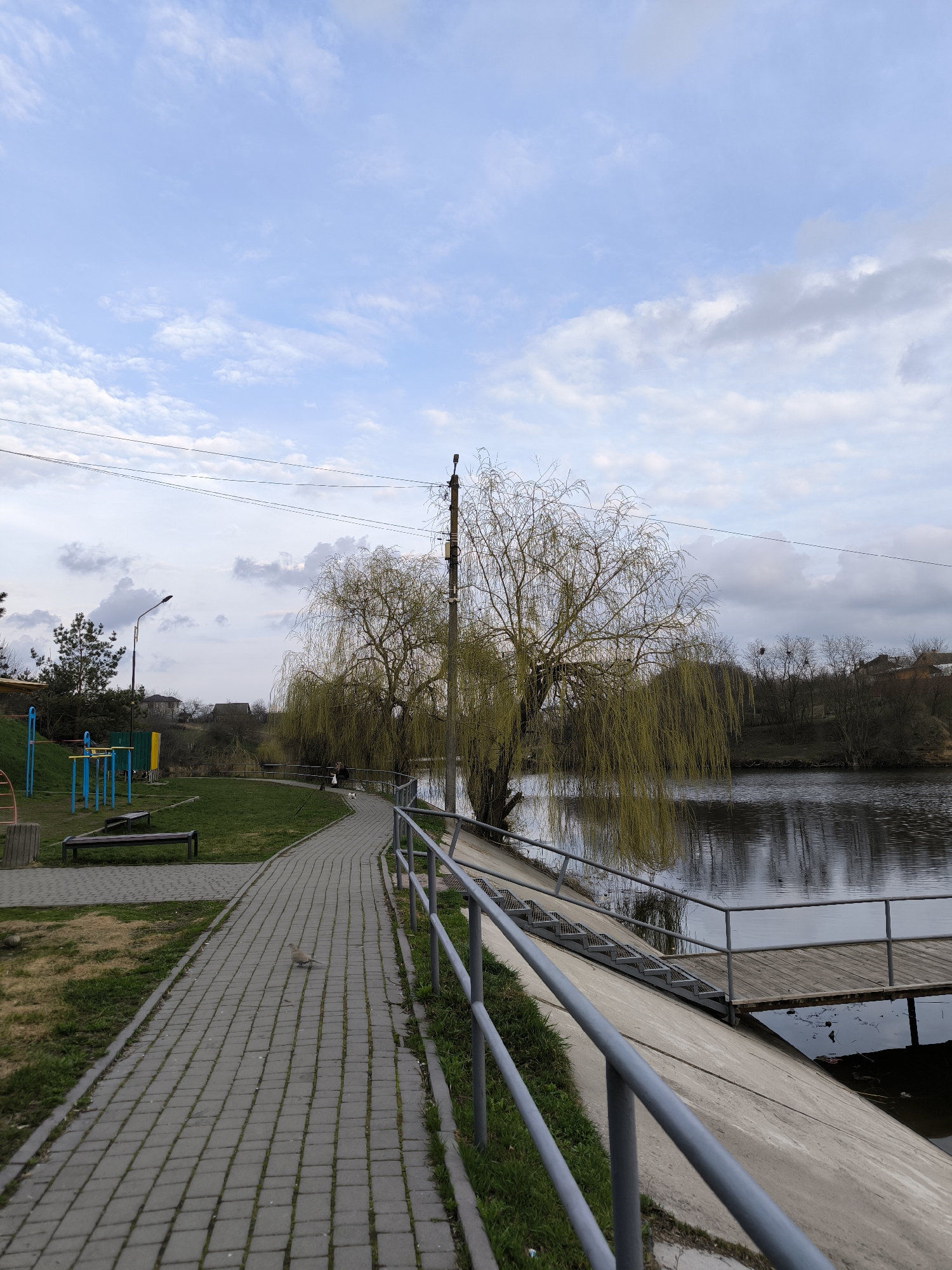 The image depicts a park in Vinnytsia, Ukraine, alongside the Southern Bug River. A paved pathway, constructed of gray rectangular stones, winds alongside a grassy area on the left and a concrete embankment with a metal railing on the right. The railing protects the edge of the embankment that slopes down to the water. A weeping willow tree stands between the path and the river. In the background, there is a playground with blue and yellow equipment and a park bench. Beyond the park area, there are residential buildings and trees on a slightly elevated terrain. The sky is partly cloudy with a mix of light blue and gray hues. The river's surface reflects the surrounding environment. A wooden platform extends into the river supported by beams. A gray dove walks along the pathway. An electrical pole stands next to the weeping willow tree.