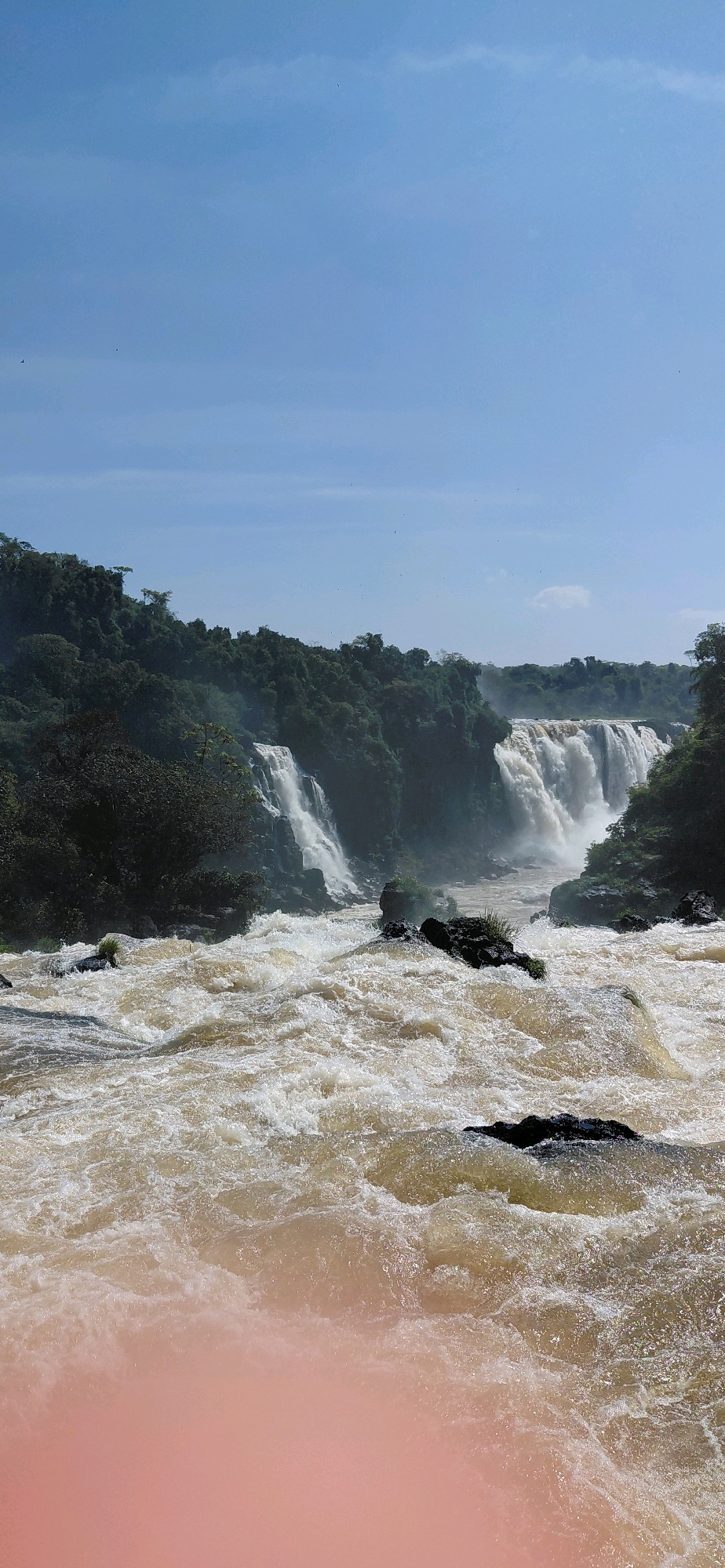 This image captures a majestic scene of the Iguaçu Falls, located in Foz do Iguaçu, Brazil. The powerful waterfalls dominate the background, with multiple cascades of milky-brown water plunging over rocky ledges amidst lush green foliage. The foreground is filled with a turbulent, churning river, its surface a chaotic mass of white foam and eddies, reflecting the bright, clear blue sky. Scattered rocks emerge from the rapids, adding to the wild and untamed nature of the scene. The sky is expansive, with a few wispy clouds, and the overall impression is one of immense natural power and beauty.