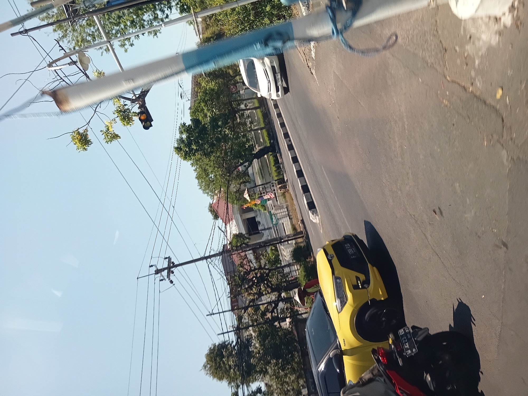 A yellow car, a white car, and a motorcycle are on a street in Surabaya, Indonesia. The yellow car is in the foreground, and the white car is in the background. The motorcycle is in the left foreground. The street is paved, and there are trees and buildings on either side. The sky is clear and blue. A person wearing a conical hat and red shirt is standing next to the yellow car. A traffic light is visible in the background.