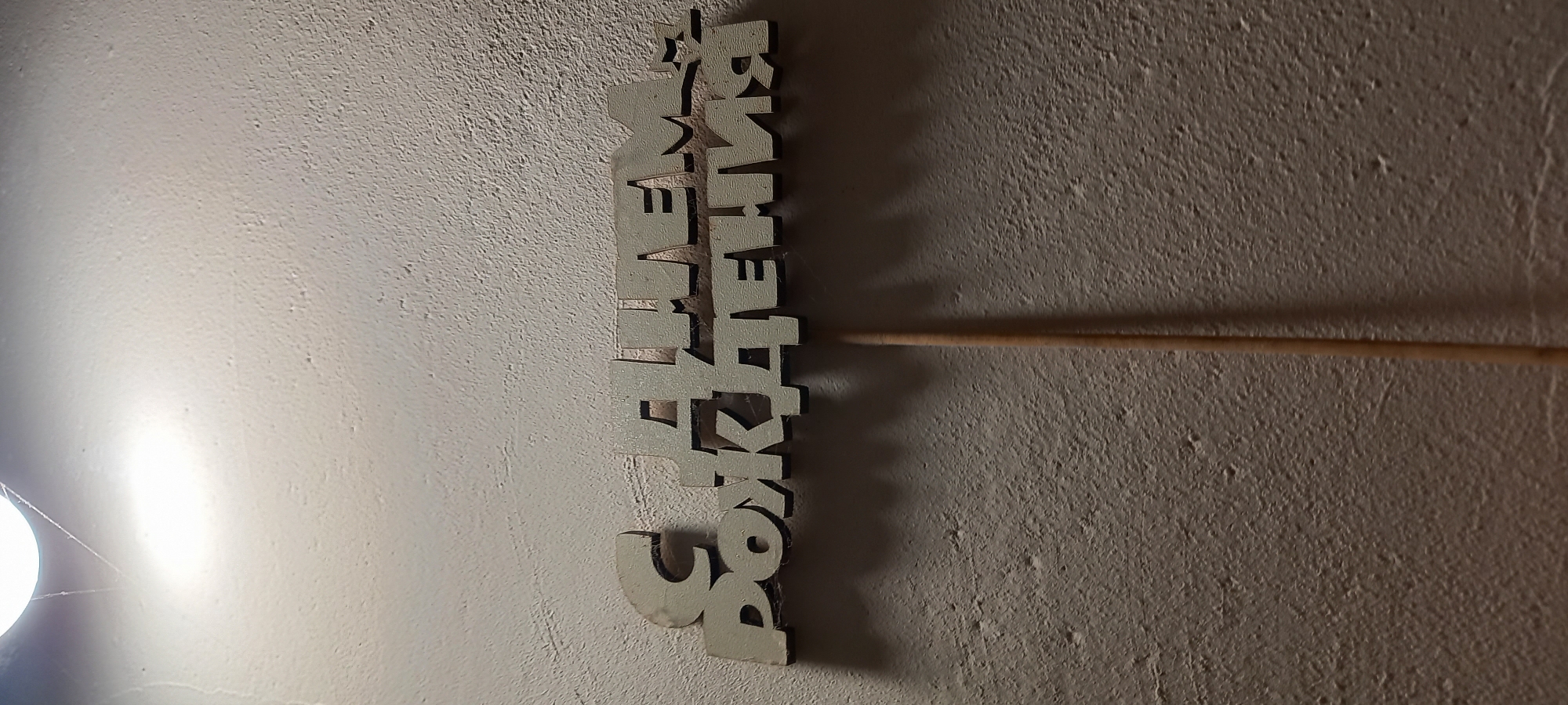 The image displays a decorative sign against a textured, light-colored wall in Kryvyi Rih, Ukraine. The sign is made of a light-colored material, likely wood or MDF, with dark edges, and spells out the Russian phrase "С ДНЁМ РОЖДЕНИЯ," which translates to "Happy Birthday." The sign is oriented vertically in the frame. A thin wooden dowel or stick extends from the right side of the sign.
A bright, artificial light source, identified in one of the crops as a bare light bulb, is visible in the lower-left portion of the image. This intense lighting casts distinct shadows to the right of the sign and stick, and strongly highlights the uneven, rough texture of the wall. The strong artificial illumination suggests an indoor setting, possibly in the evening or at night. The presence of the "Happy Birthday" sign indicates a celebratory event, likely a birthday, is either underway or being prepared.