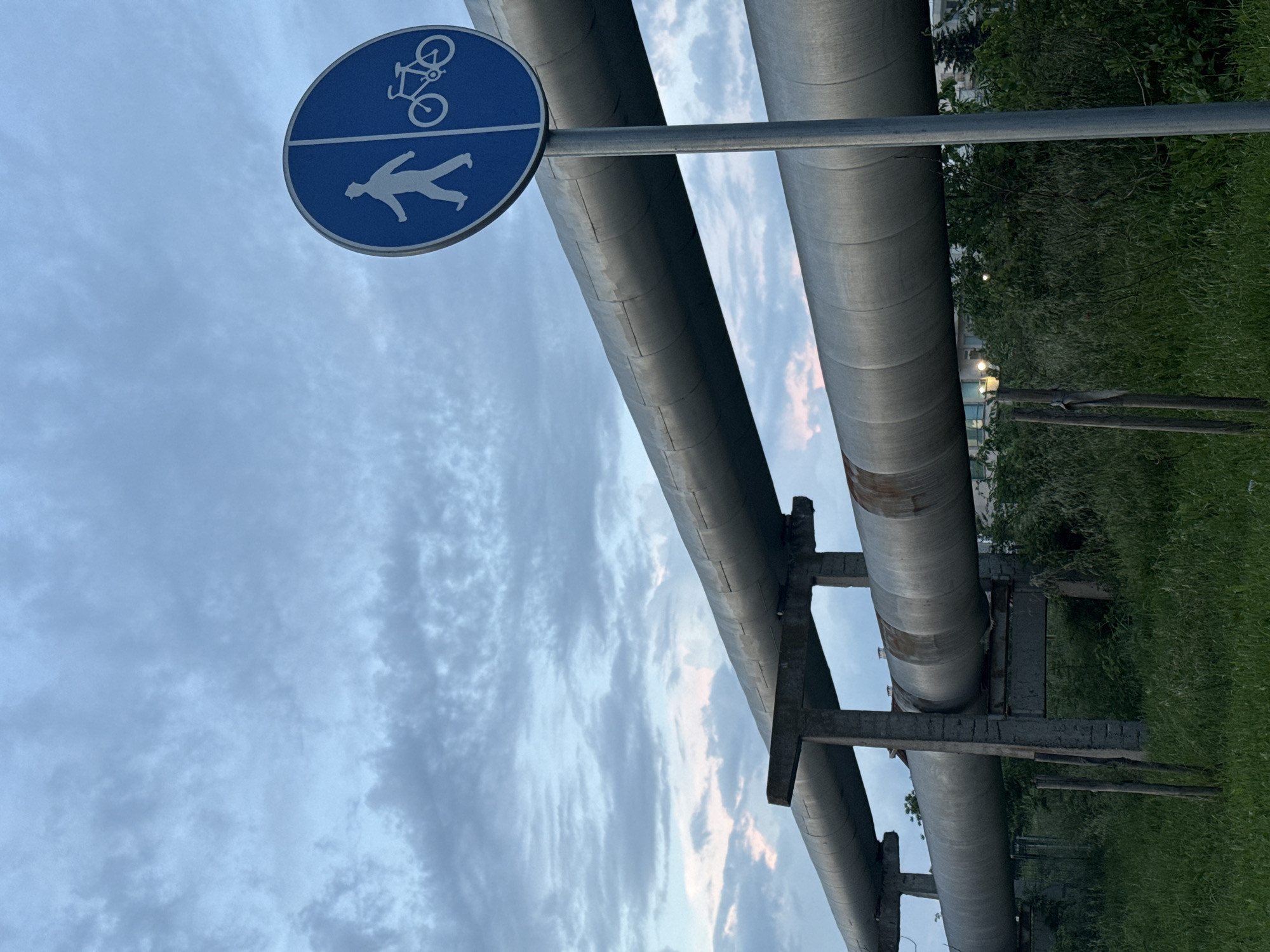 A low-angle, outdoor shot presents a pathway designated for both pedestrians and cyclists, as indicated by a blue sign with white icons. The sign is positioned adjacent to two parallel, elevated metal pipes that appear to be part of an industrial or utility system. The pipes are supported by concrete or metal structures. The background reveals a blend of urban and natural elements, including greenery (likely grass and foliage) and glimpses of a building in the distance. The sky, visible above and between the pipes, is overcast with varied cloud formations. The image does not show Burgas, Bulgaria. The photo's orientation is tilted, giving a slightly skewed perspective of the scene. There are no people present in the image.